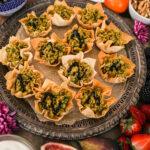 A tray of baked pastry shells filled with minced chicken and topped with a drizzle of a dark dressing surrounded by fruits and nuts.