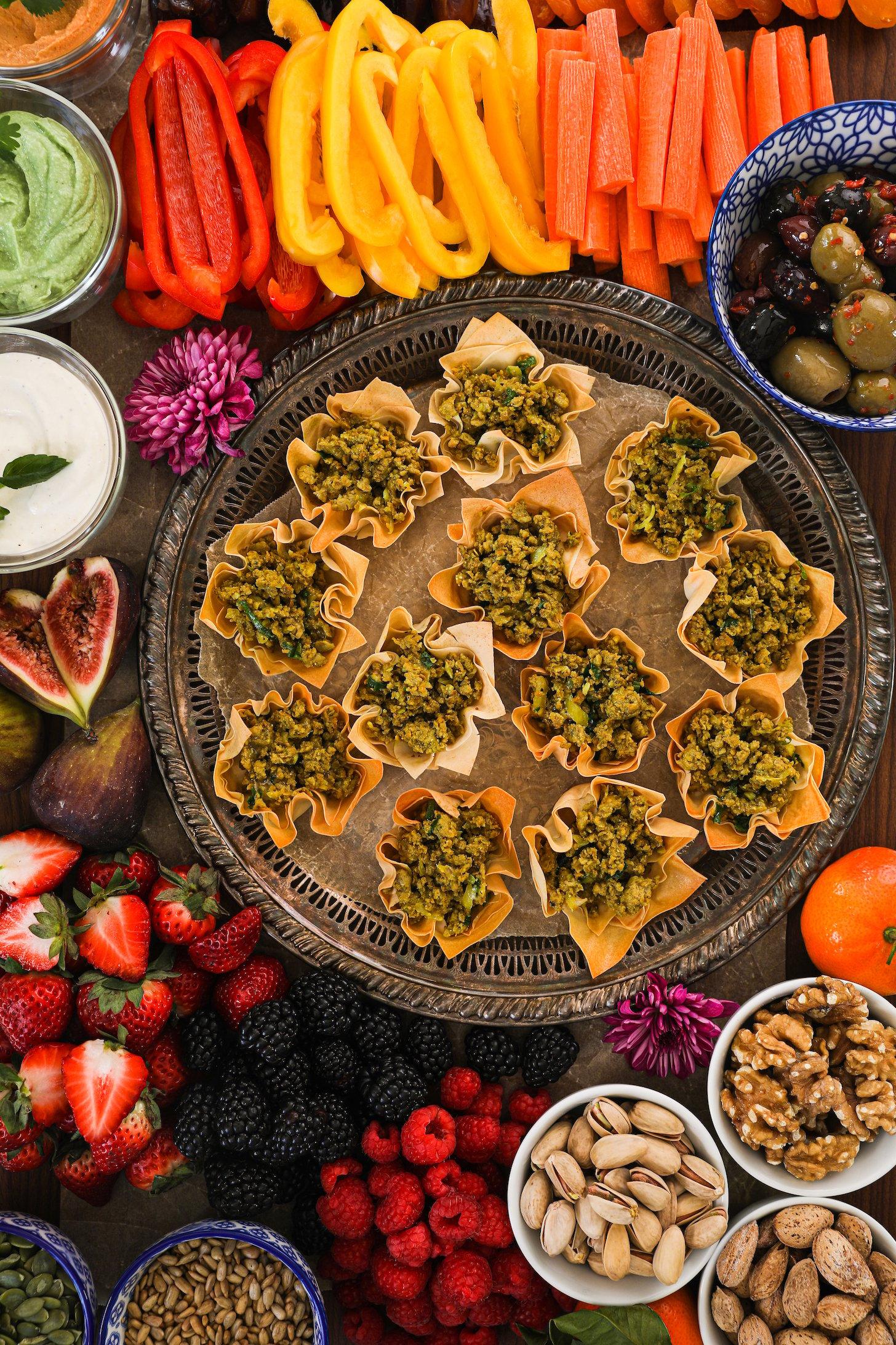 A beautiful and colourful display of food with a tray of baked pastry shells filled with chicken, fruits, veggies, nuts, olives, seeds and dips.