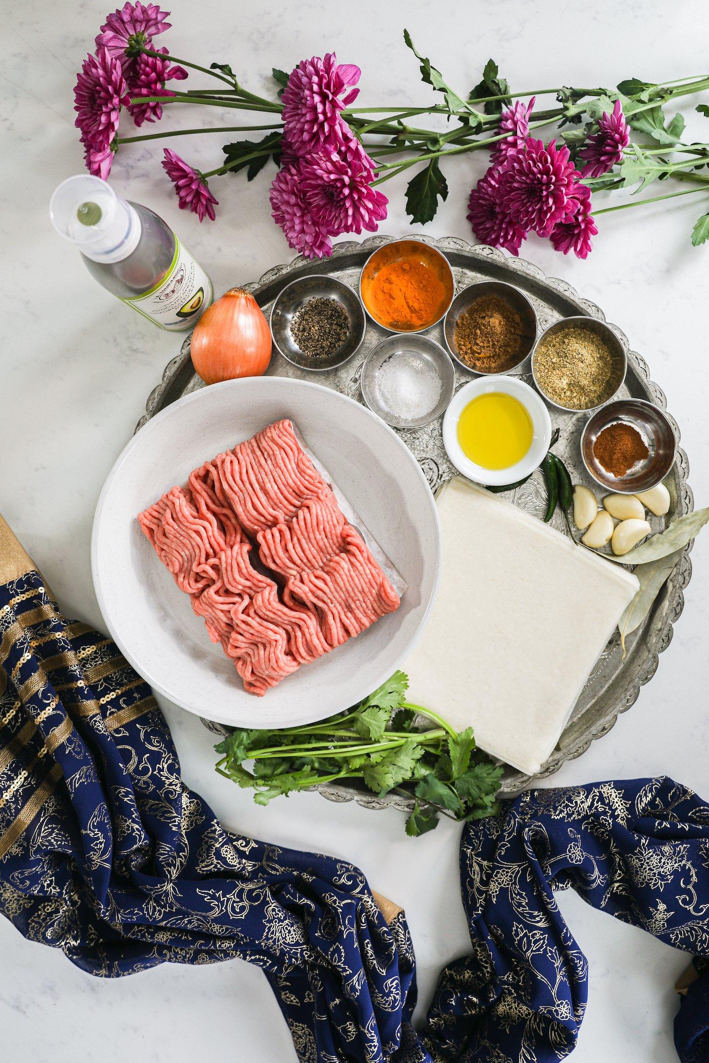 Food ingredients like minced chicken, spices, aromatics and herbs styled with pink flowers and a traditional scarf.