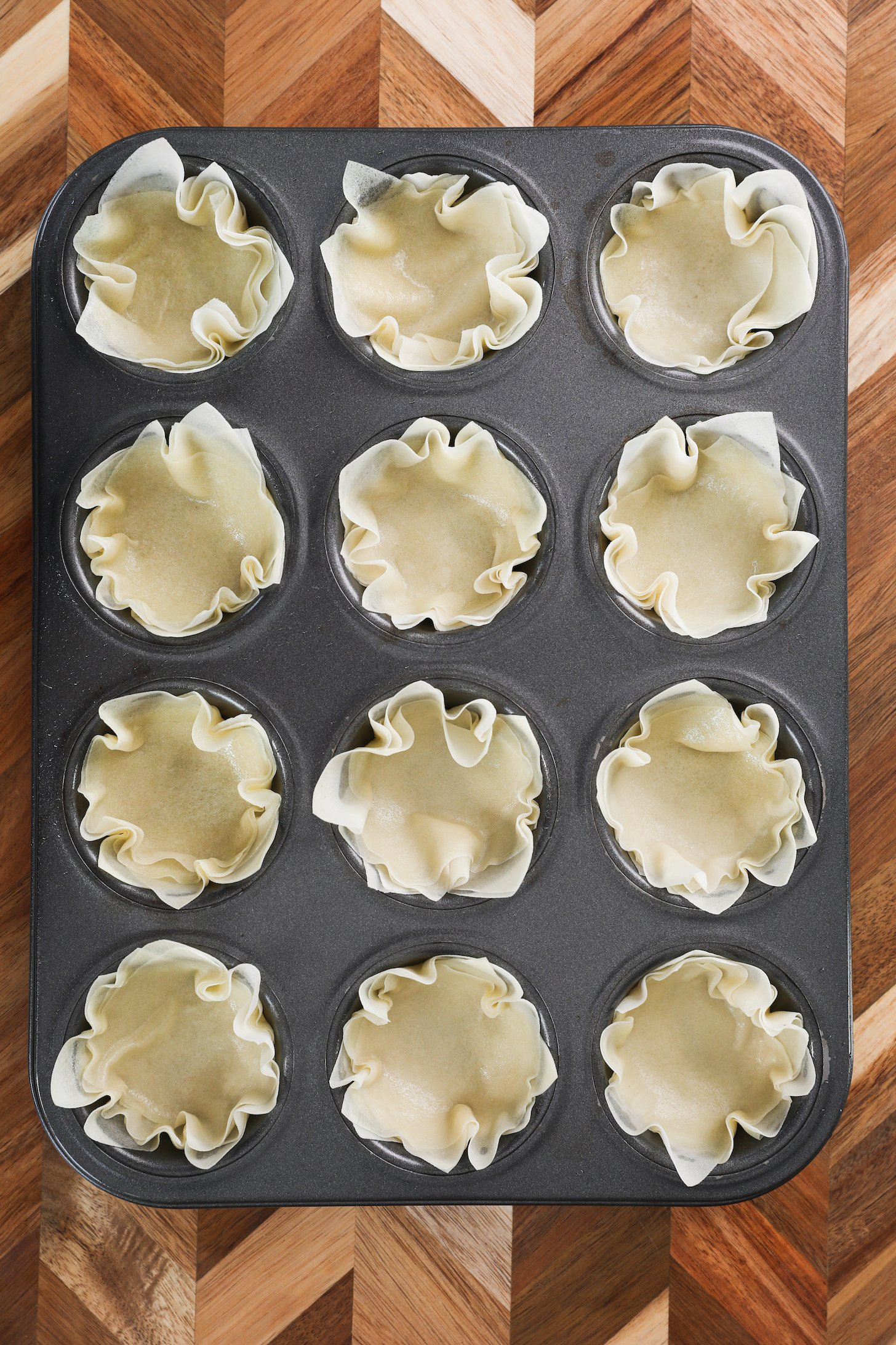 A cup cake tray holding 12 uncooked pastry shells shaped like a flower.