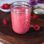 A mason jar of red sauce or dressing on a wooden board surrounded by scattered raspberries and flowers.