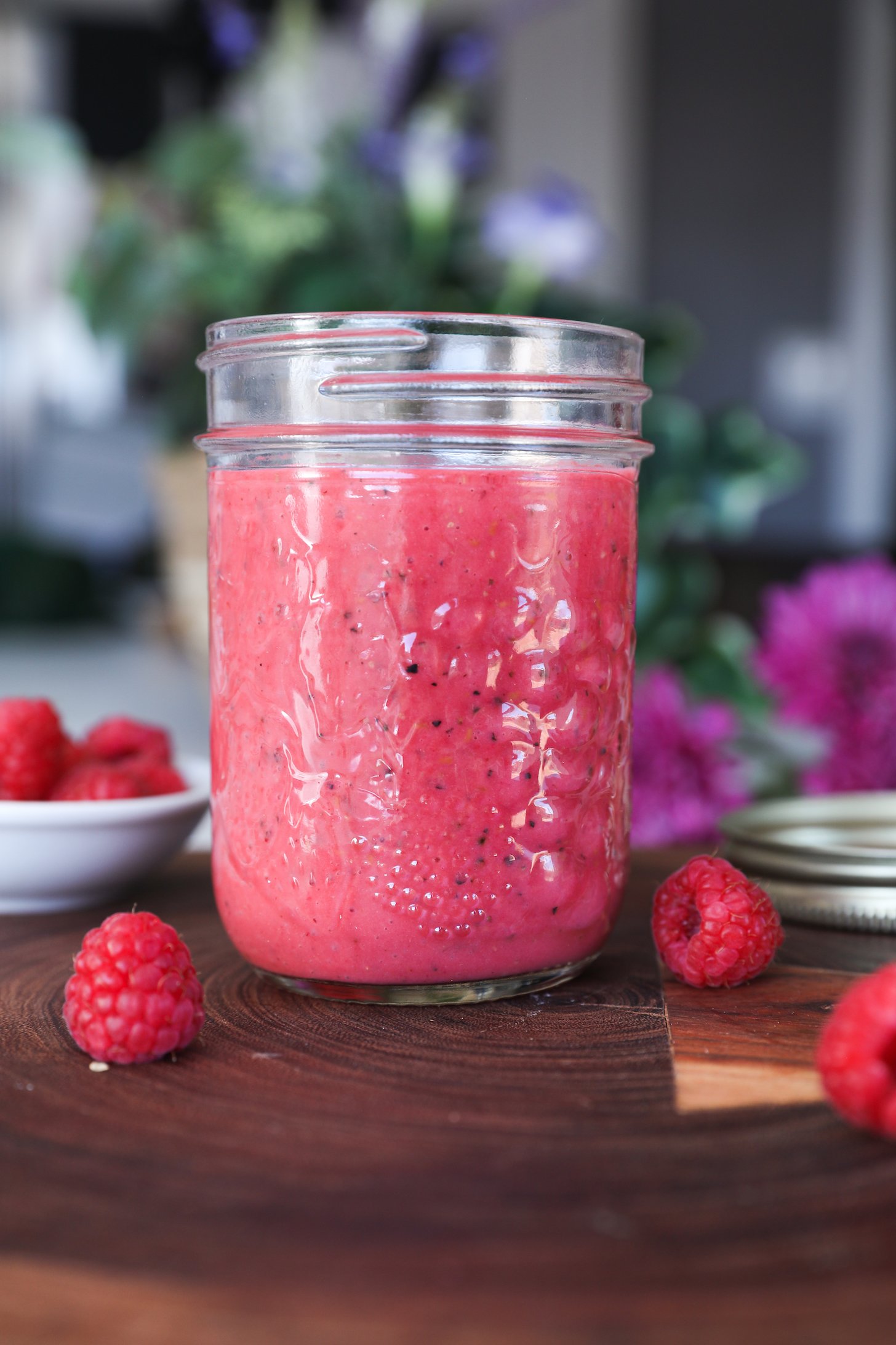 A perspective image of a mason jar of red sauce or dressing on a brown surface with several raspberries nearby.