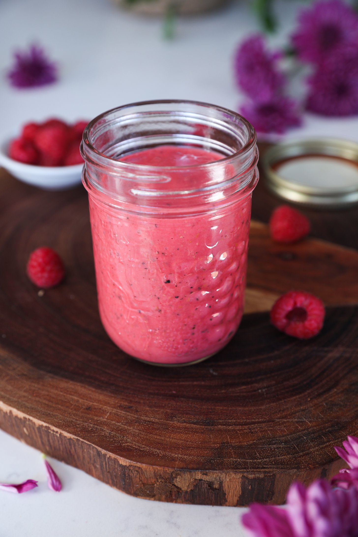 A mason jar of red sauce or dressing on a wooden board surrounded by scattered raspberries and flowers.
