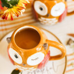 A perspective image of two owl mugs filled with chai apple cider on a striped orange mat.