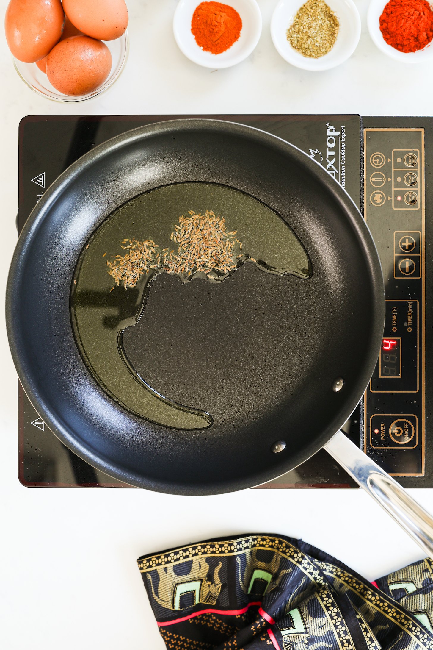 A pan with cumin frying in oil on a mobile cooktop with eggs and powdered spices.