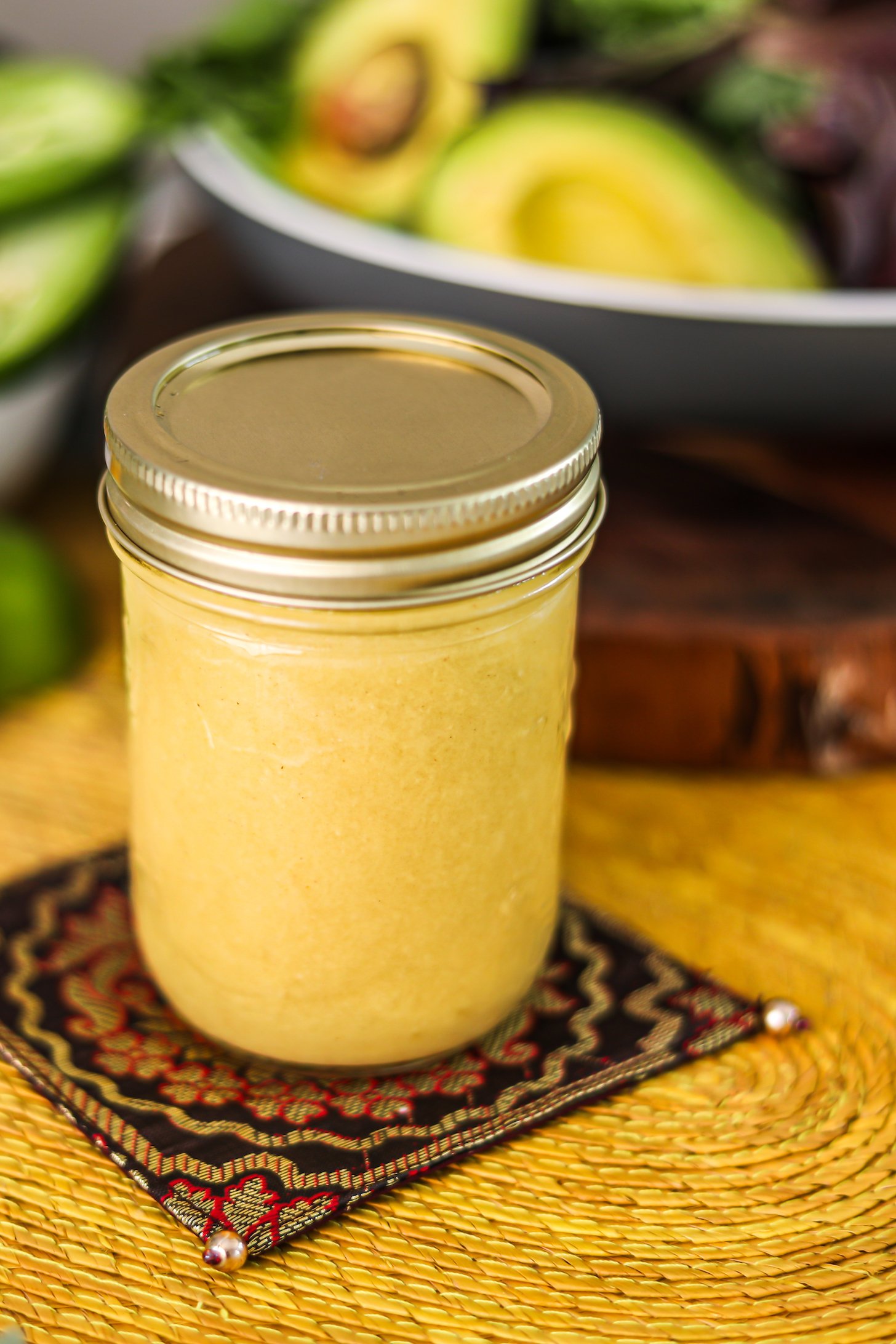 Perspective image of a mason jar of honey mustard dressing on a decorative mat with avocados in the bacground.