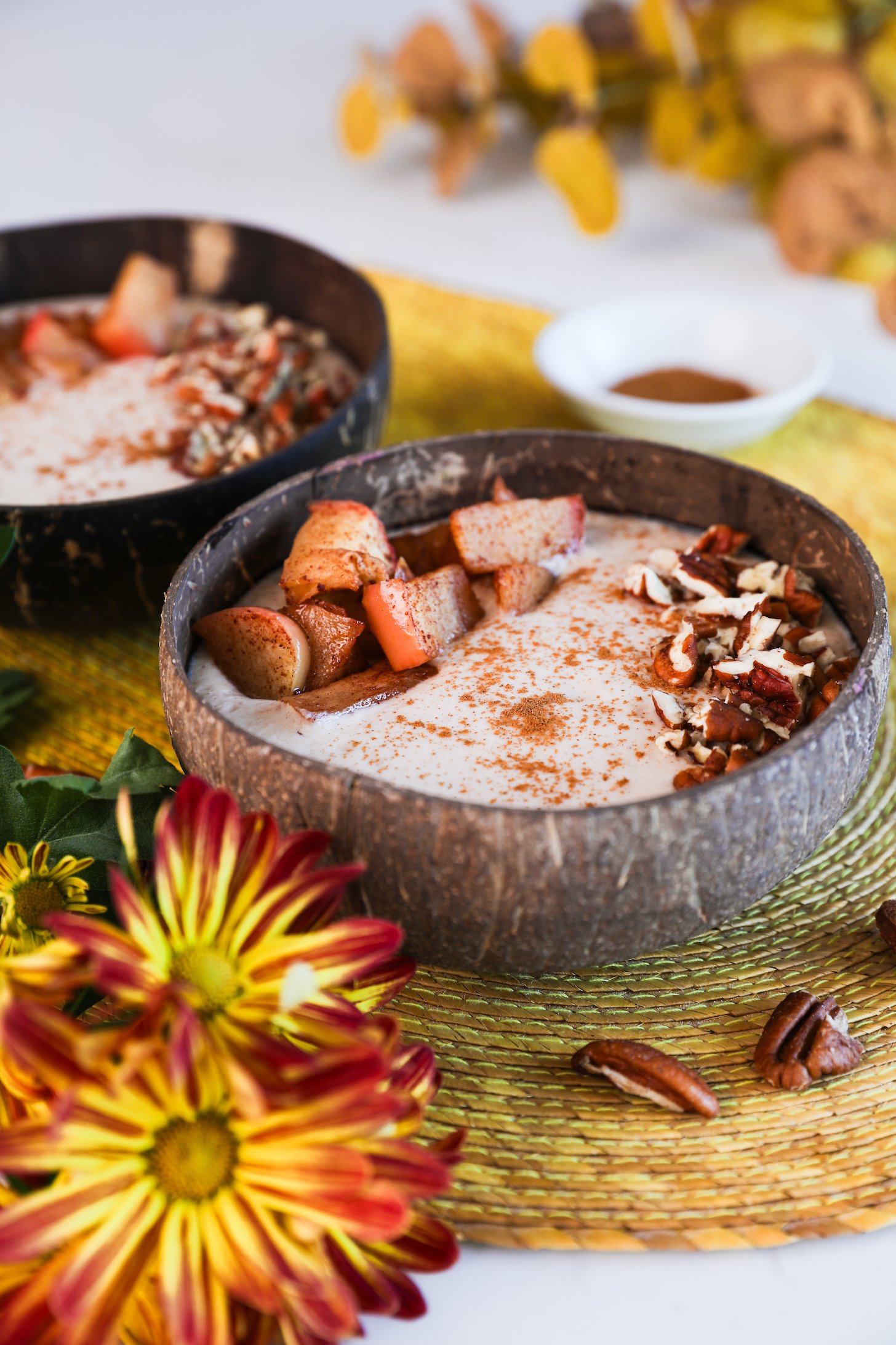 Image of two coconut bowls filled with beige smoothie, garnished with baked apple slices, pecans, and cinnamon, with bright yellow flowers in the foreground.