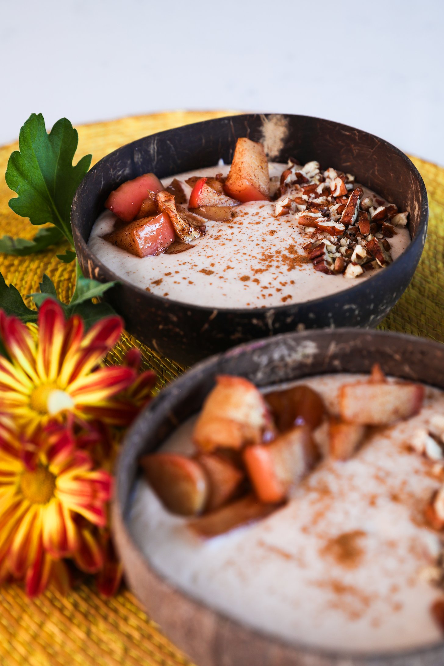 Two coconut bowls with beige smoothie, garnished with baked apple slices, pecans, and cinnamon, one in focus, the other blurred, with nearby bright yellow flowers.