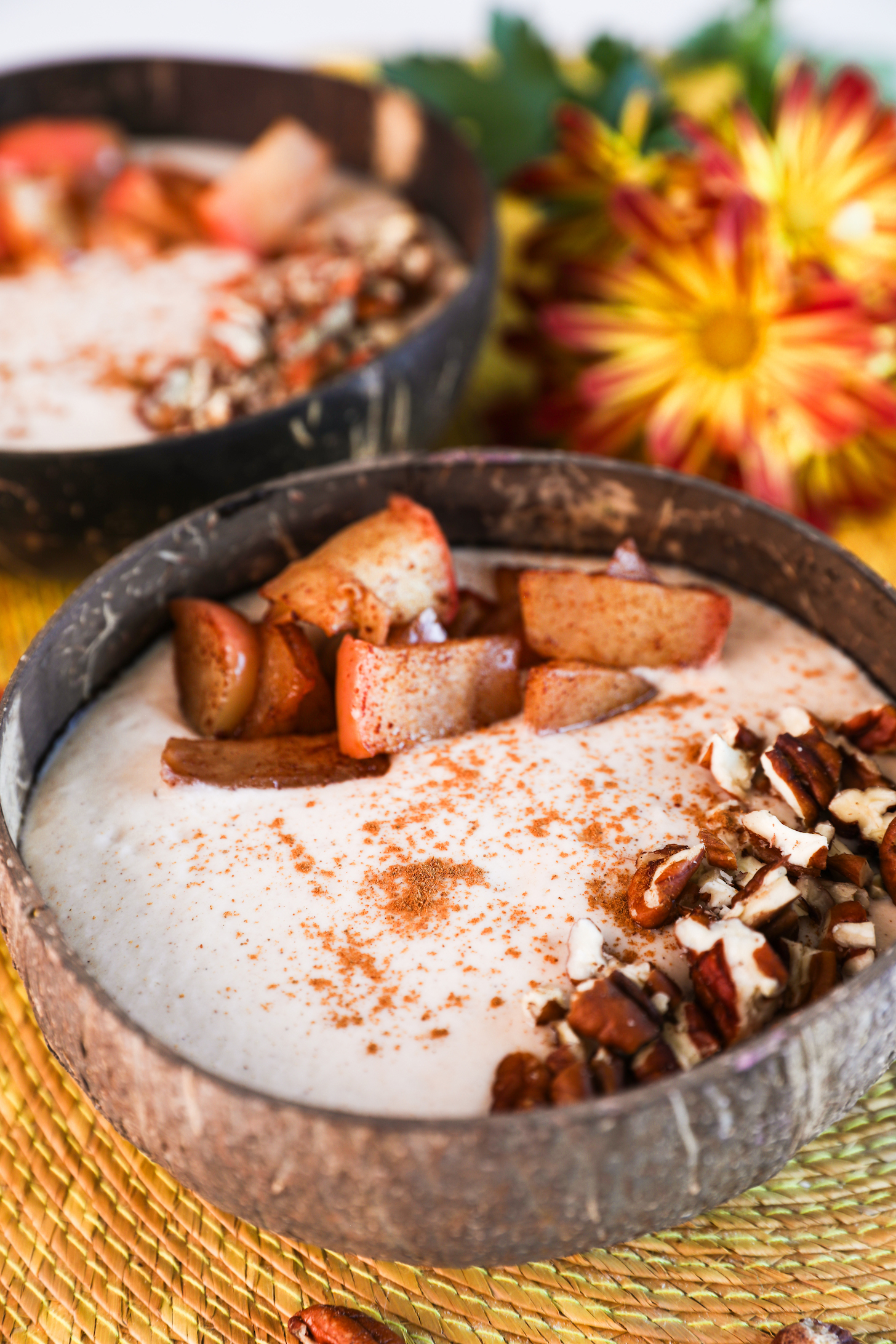 Close up image of two coconut bowls with beige smoothie, garnished with baked apple slices, pecans, and cinnamon, one in focus, the other blurred, with nearby bright yellow flowers.