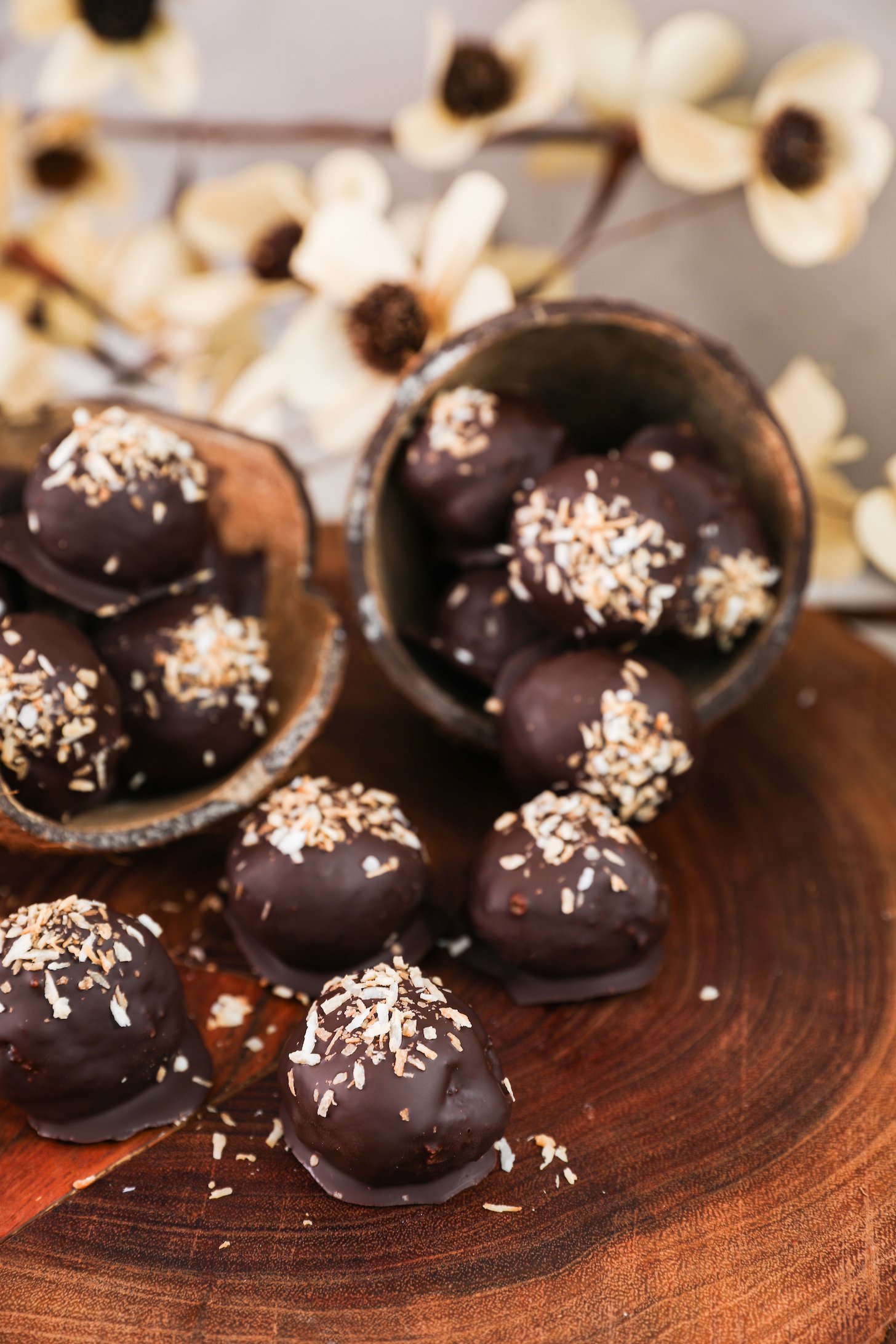 Close up image of chocolate-covered balls with a coconut topping displayed in coconut shells, set against a backdrop of beige flowers.