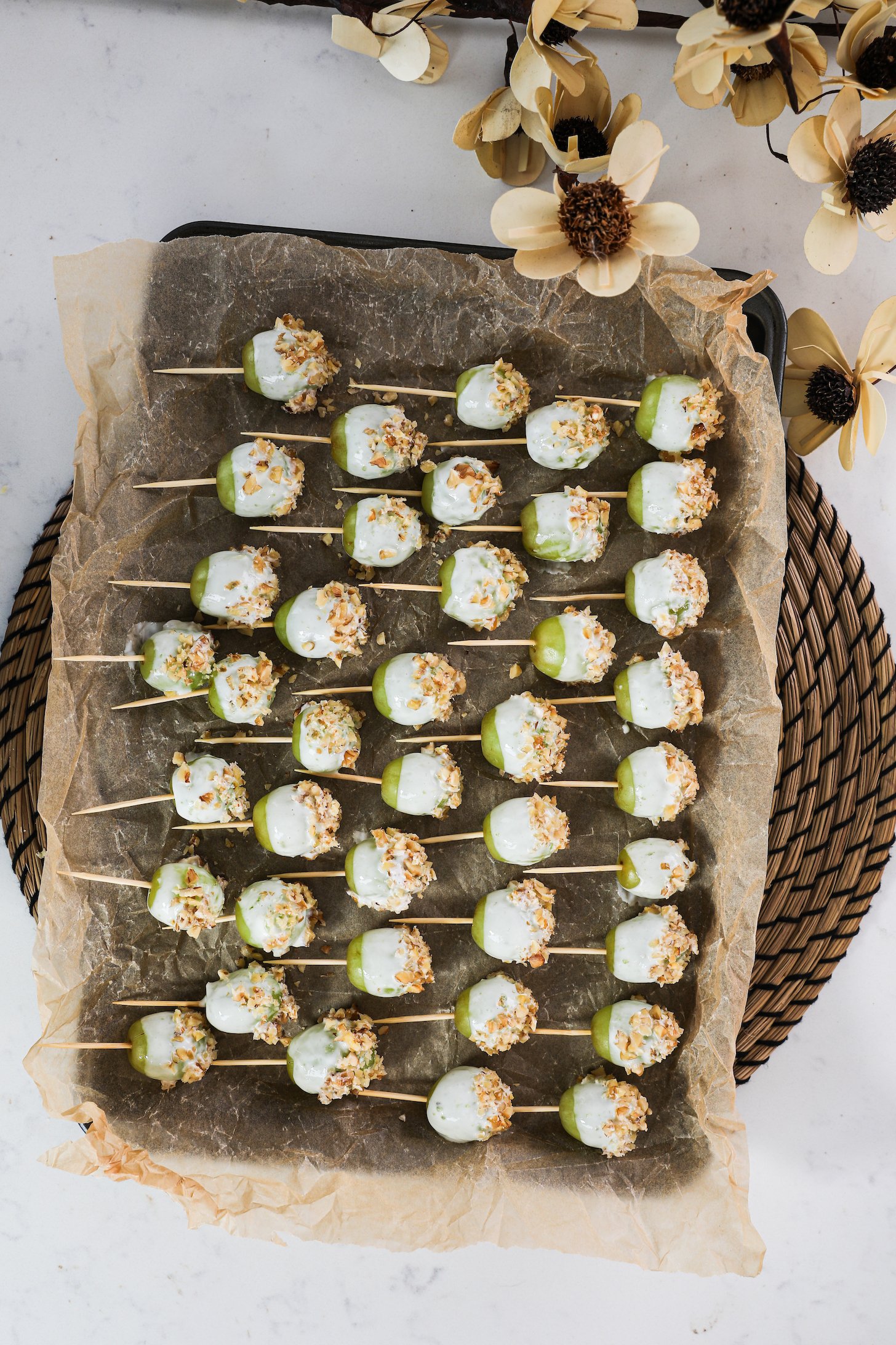 A meticulously arranged baking sheet showcases unfrozen green grapes, each on a wooden pick, elegantly coated in yogurt and sprinkled with crushed nuts.