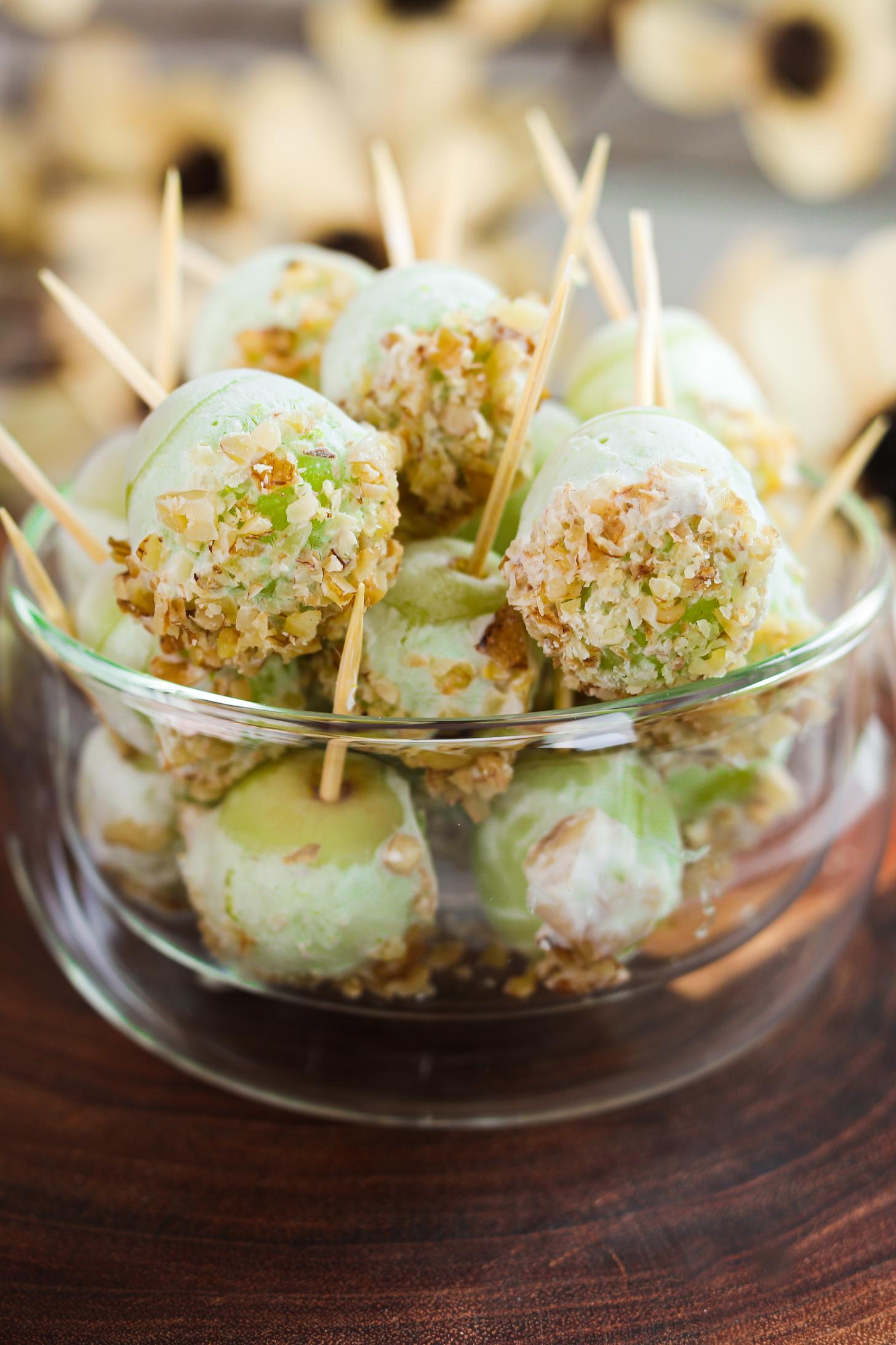 A perspective close up image displays a bowl filled with grapes on wooden picks, coated in yogurt, and garnished with crushed nuts.