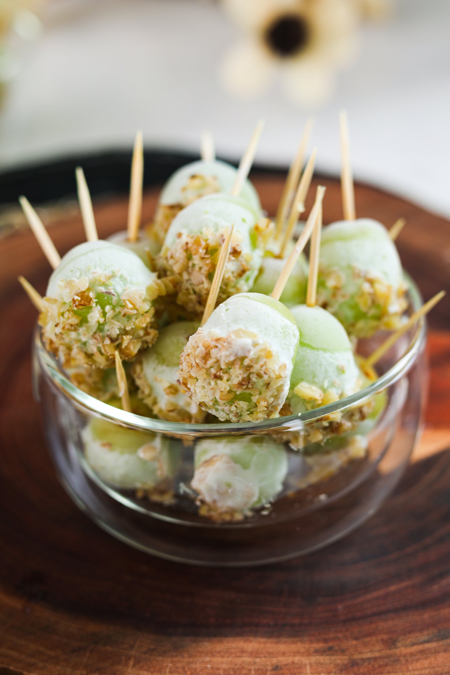 An image displays a bowl filled with grapes on wooden picks, coated in yogurt, and garnished with crushed nuts.