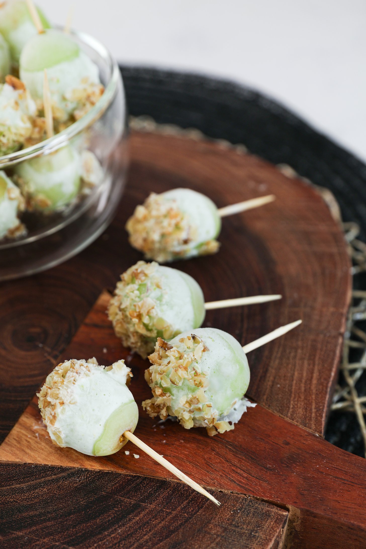 An image displays four grapes on wooden picks, covered in yogurt and topped with crushed nuts.