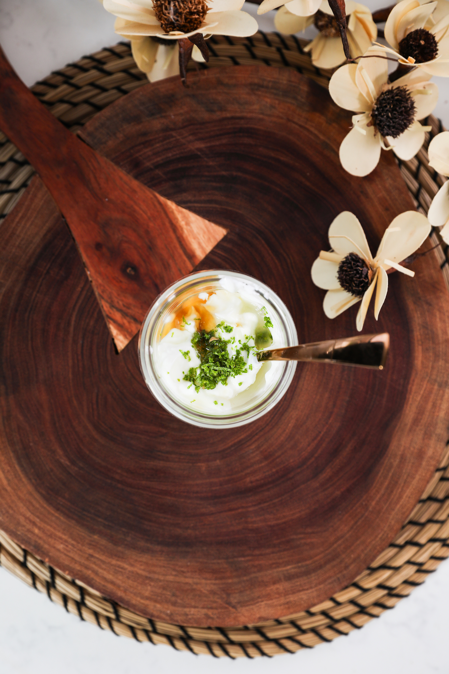 A jar of yogurt with lime zest and maple syrup on top, plus a spoon inside, all on a wooden board with wooden flowers nearby.