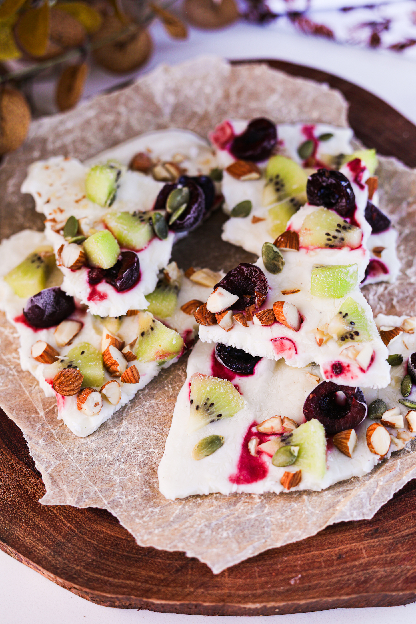 Perspective image featuring stacked frozen yogurt bark pieces, topped with fruits and nuts styled on a wooden serving board.