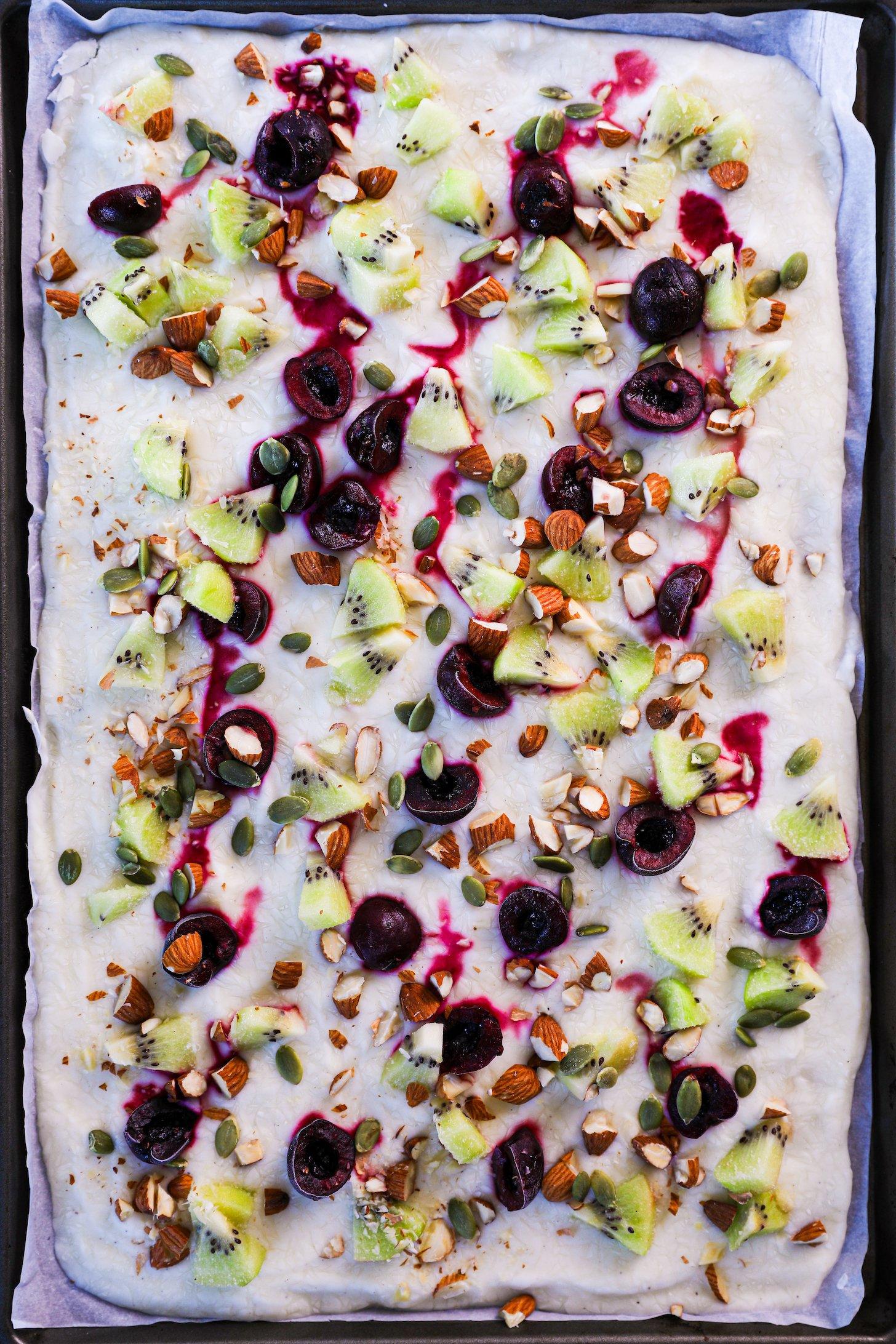 A smooth layer of frozen yogurt garnished with a variety of diced fruits and nuts, showcased on a tray with raised edges and parchment paper lining.