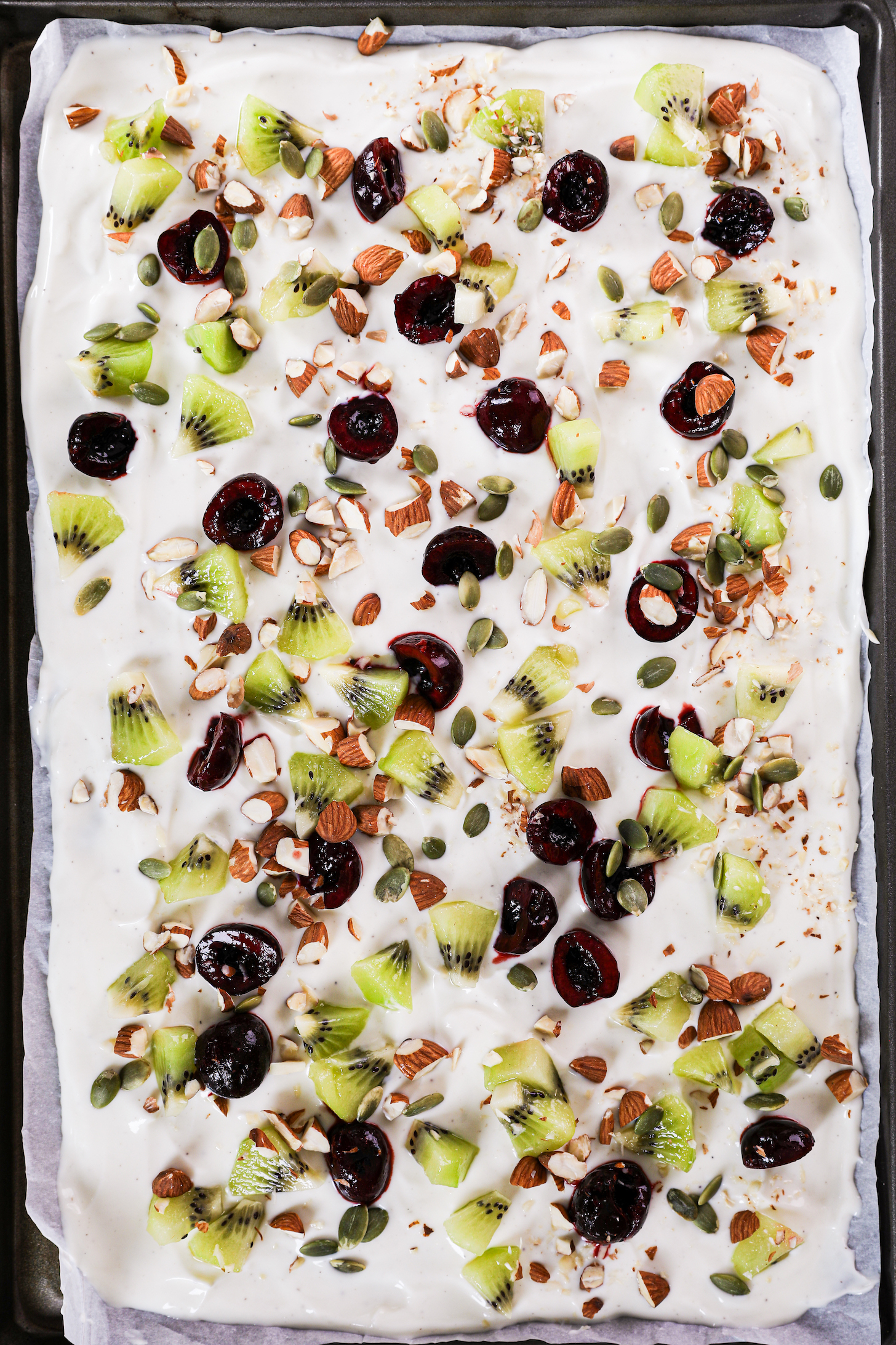 A smooth layer of yogurt garnished with a variety of diced fruits and nuts, showcased on a tray with raised edges and parchment paper lining.