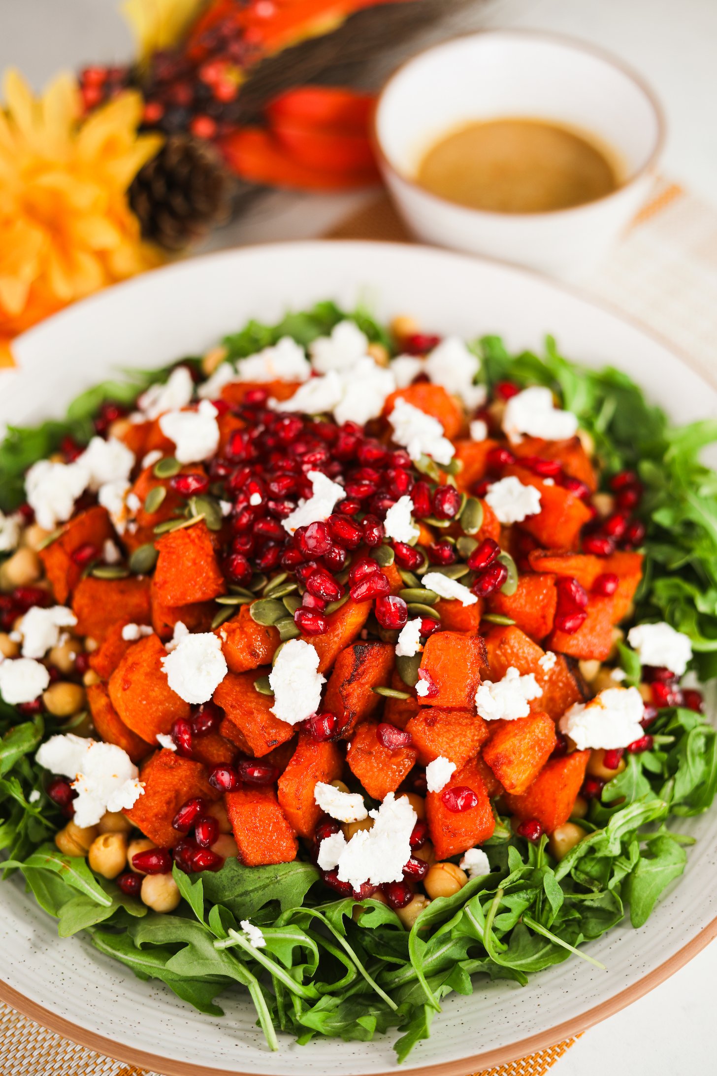 Perspective image showing a layered salad with a bed of arugula, roasted pumpkin pieces, pomegranate kernels, seeds, and chunks of goat cheese on top.