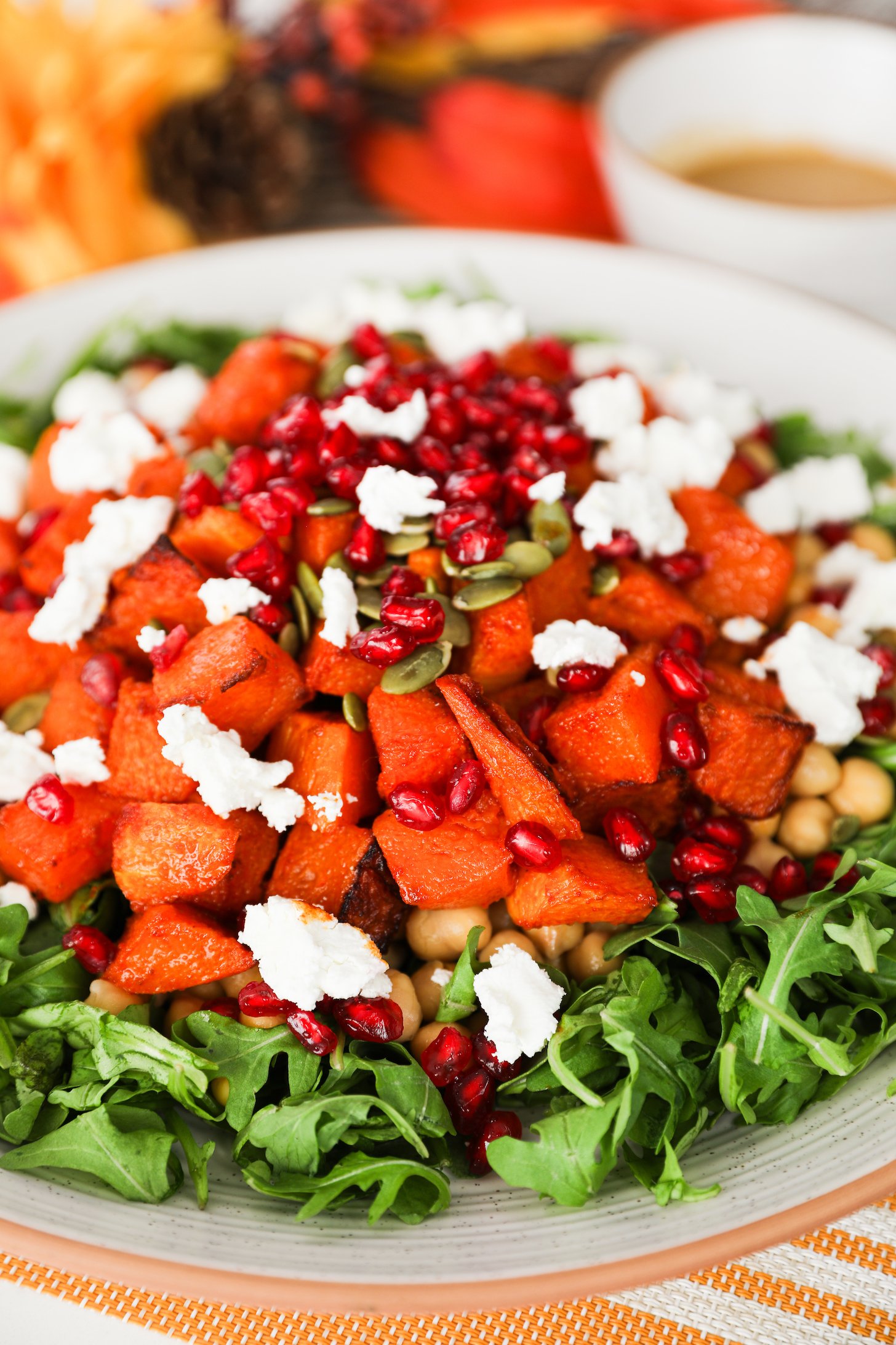 A close-up image featuring a plate of arugula topped with roasted pumpkin pieces, pomegranate kernels, seeds, and chunks of goat cheese.