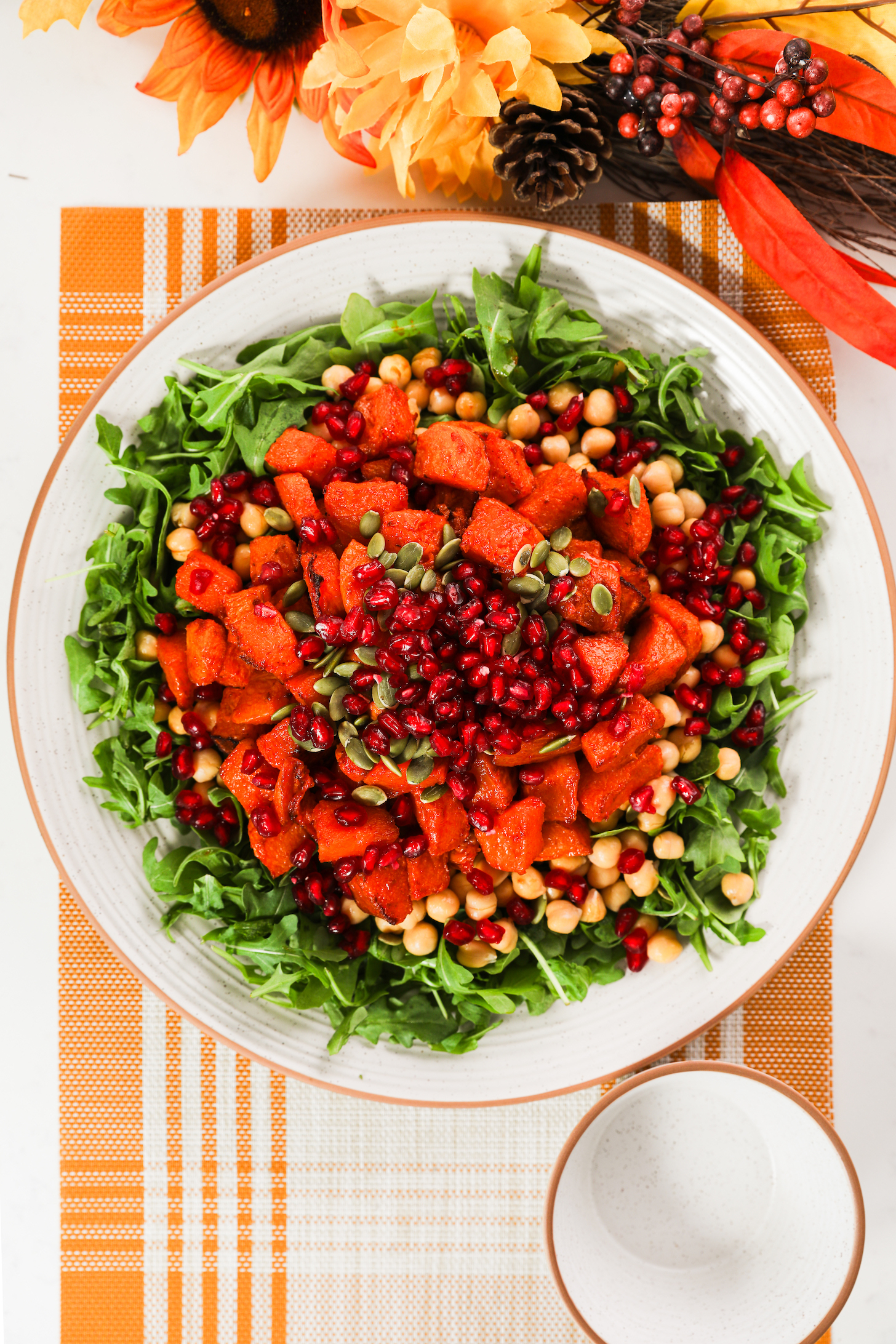 A large plate with a bed of arugula, chickpeas, roasted pumpkin pieces, pomegranate kernels and pumpkin seeds set on a placemat with orange and yellow flowers nearby.