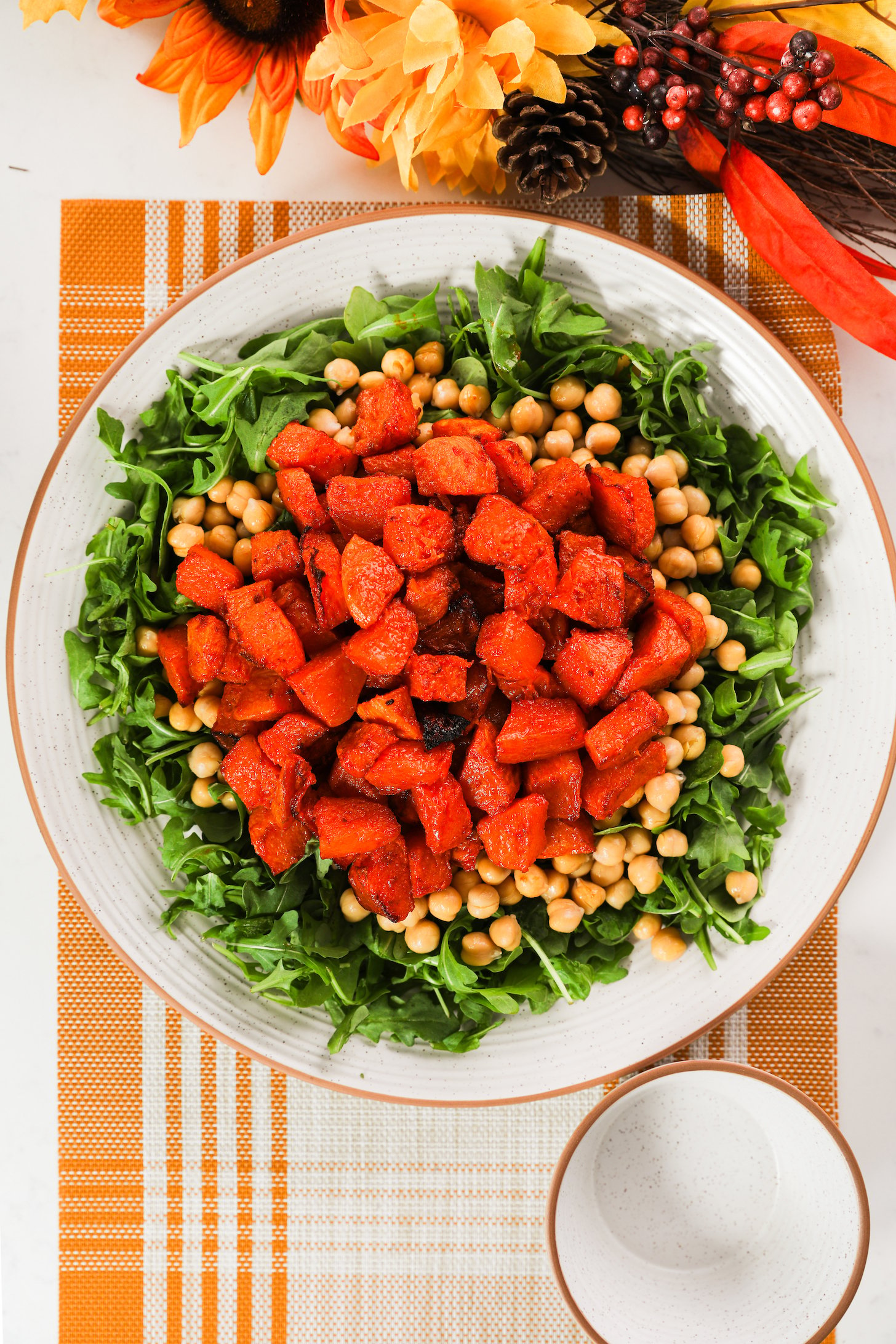 A large plate with a bed of arugula, chickpeas, and roasted pumpkin pieces, set on a placemat with orange and yellow flowers nearby.