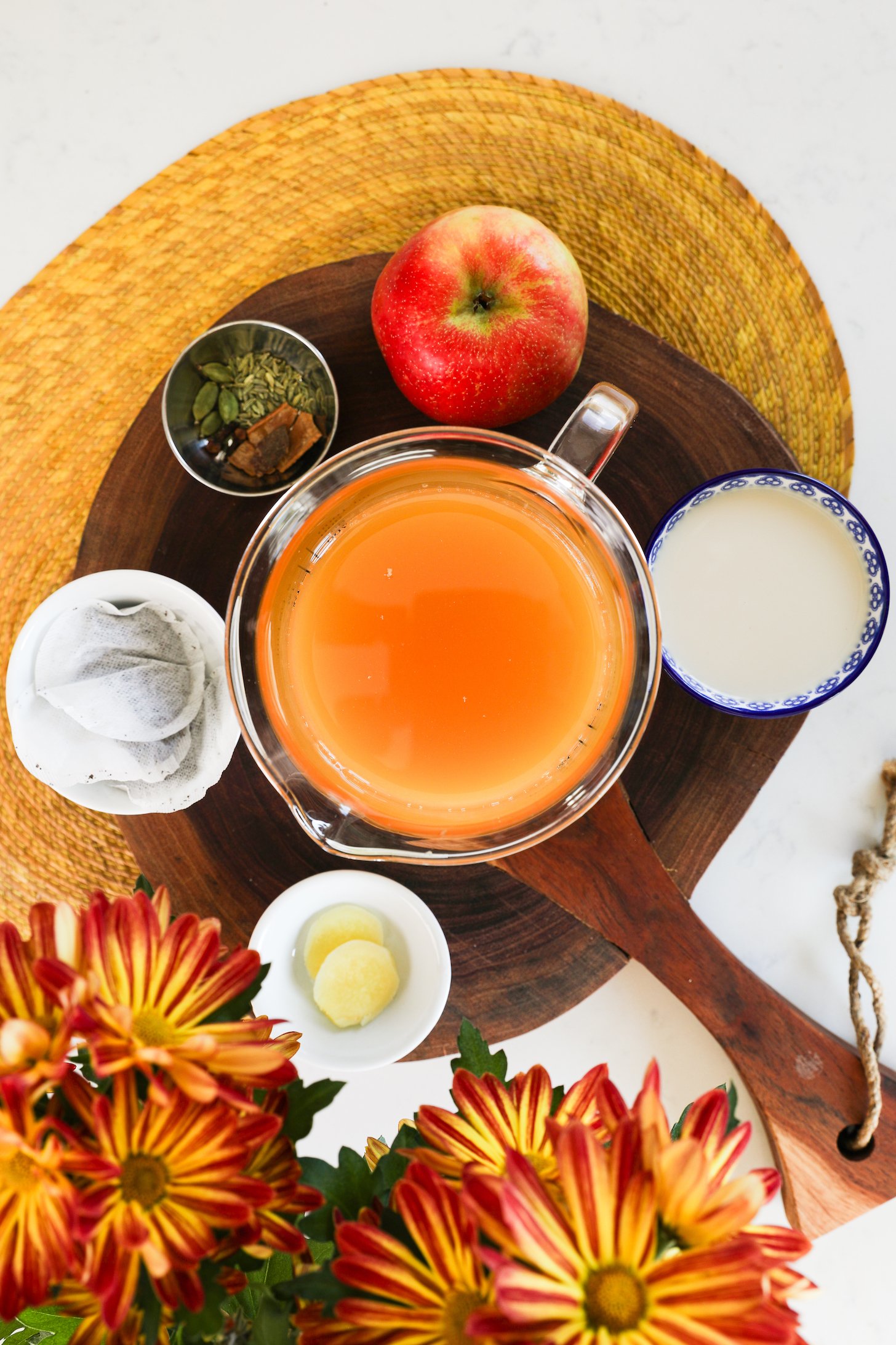 Styled food ingredients on a wooden board: apple cider, tea bags, milk, apples, and spices.
