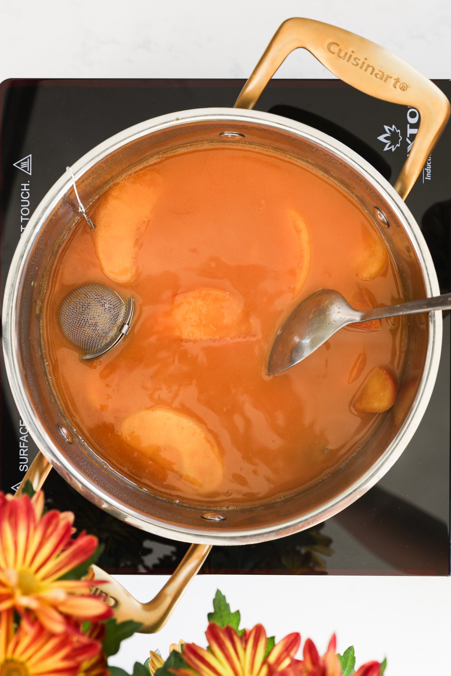 A cooking pot on a mobile cooktop holds apple cider and apple slices, with a stainless steel spice infuser and a submerged spoon.