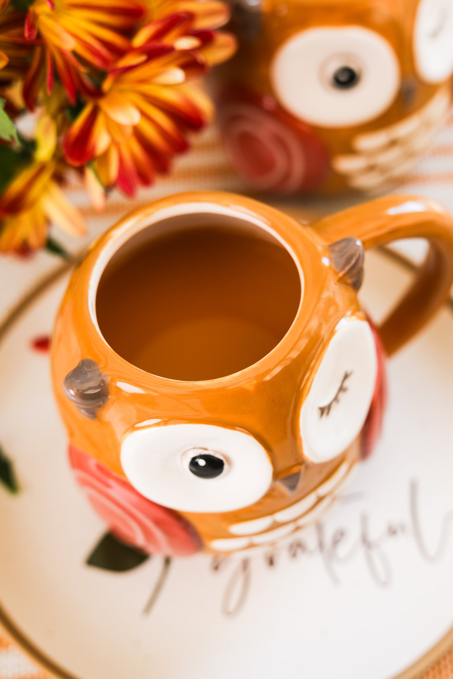 An owl mug filled with chai apple cider rests on a 'grateful' scribed plate against a backdrop of flowers.