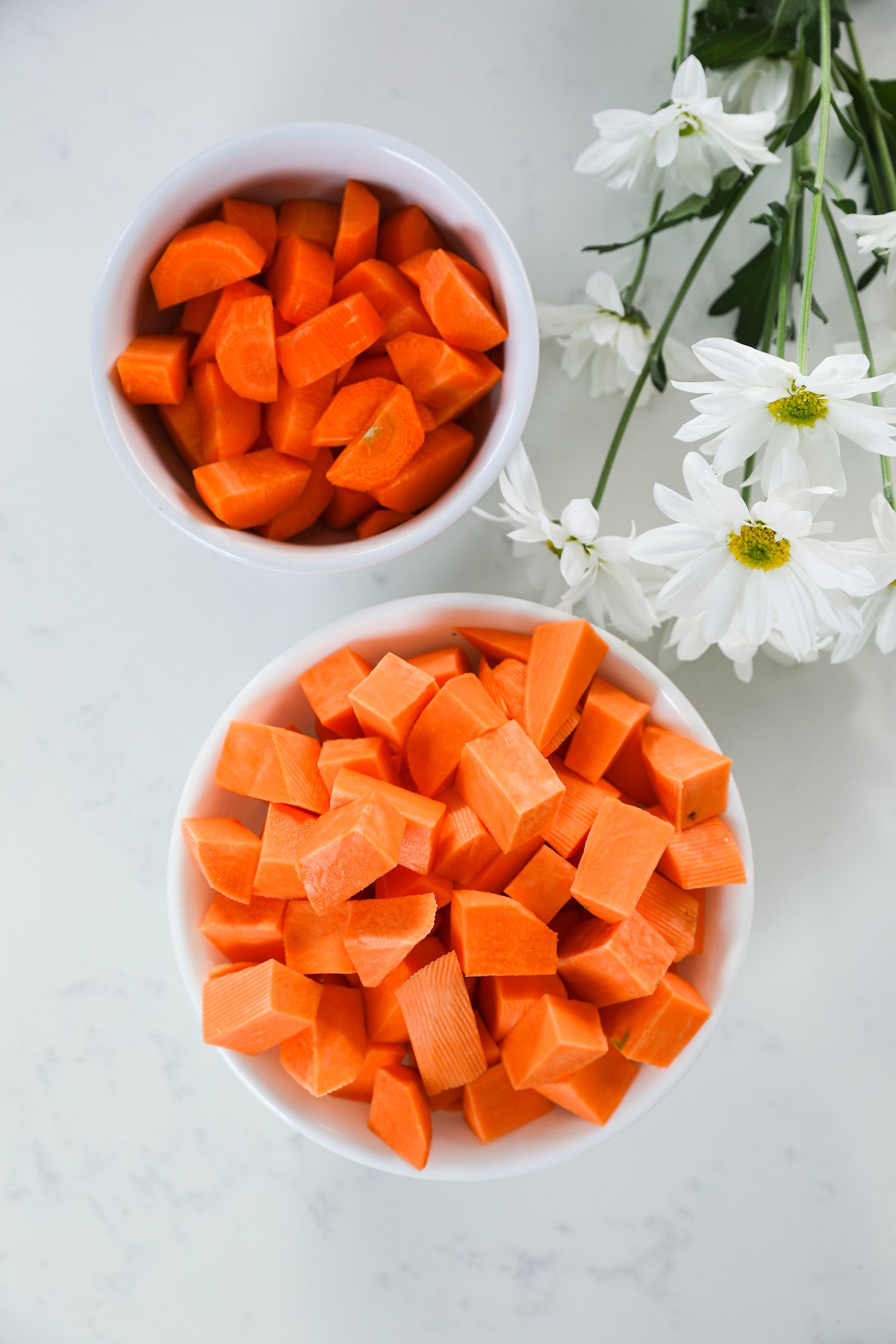 Two bowls containing chopped sweet potato and carrots, placed beside white flowers.