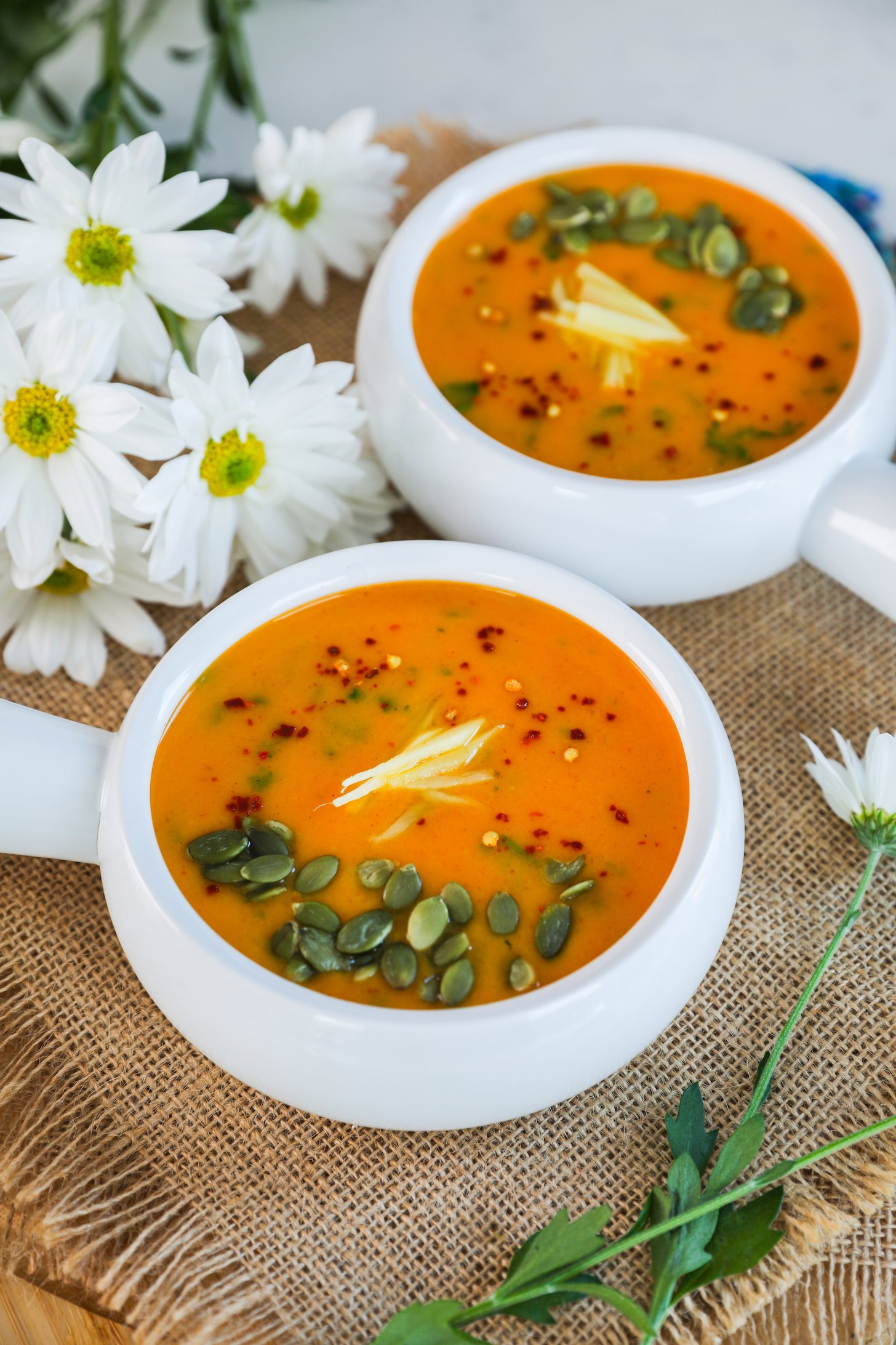 A perspective shot capturing two bowls of vibrant orange-hued soup adorned with seeds, ginger strips, and chilli flakes, complemented by white flowers in the background.