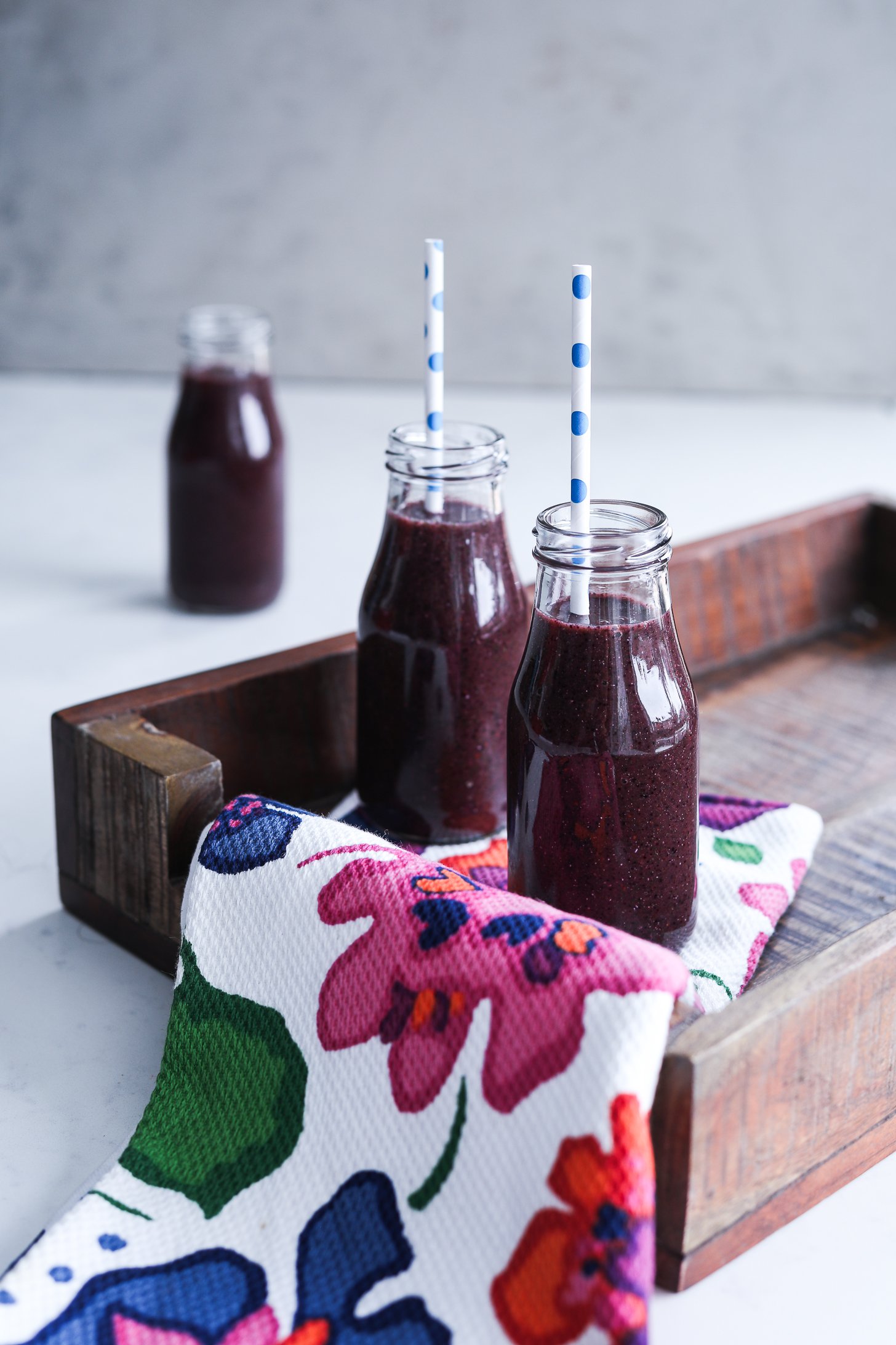 Angled perspective image of blueberry kale smoothie in small bottles with straws on a tray, covered by a cloth extending out.