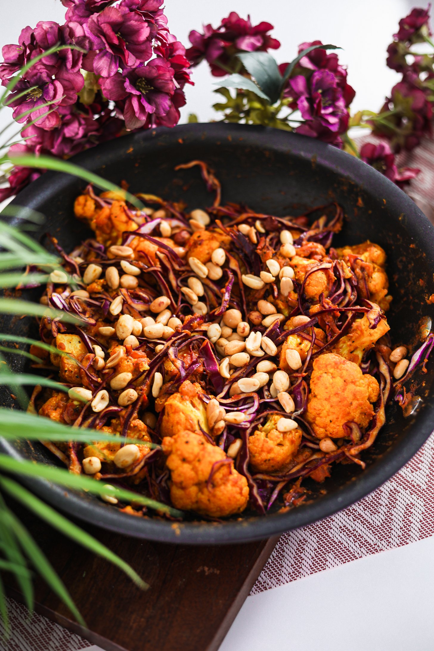 Angled photo of a pan of stir fried cabbage and cauliflower topped with peanuts with plant and flowers in the fore-and background.