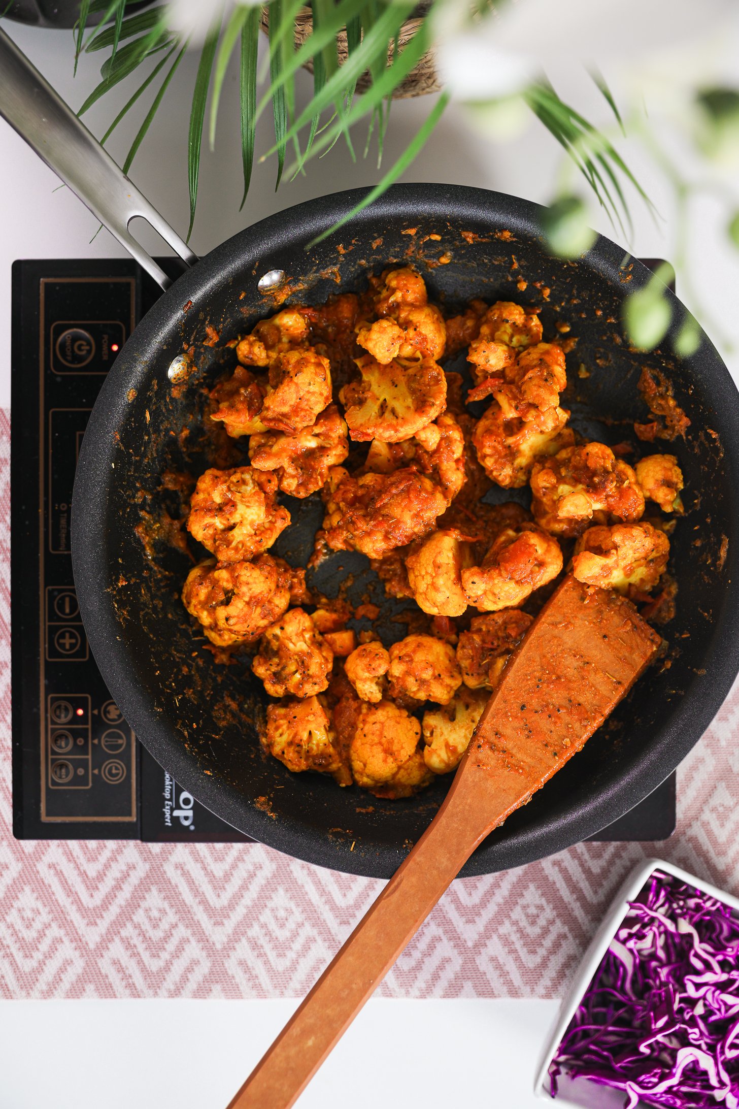Pan of spicy stir-fried cauliflower on a mobile worktop, featuring a wooden spoon within the pan.