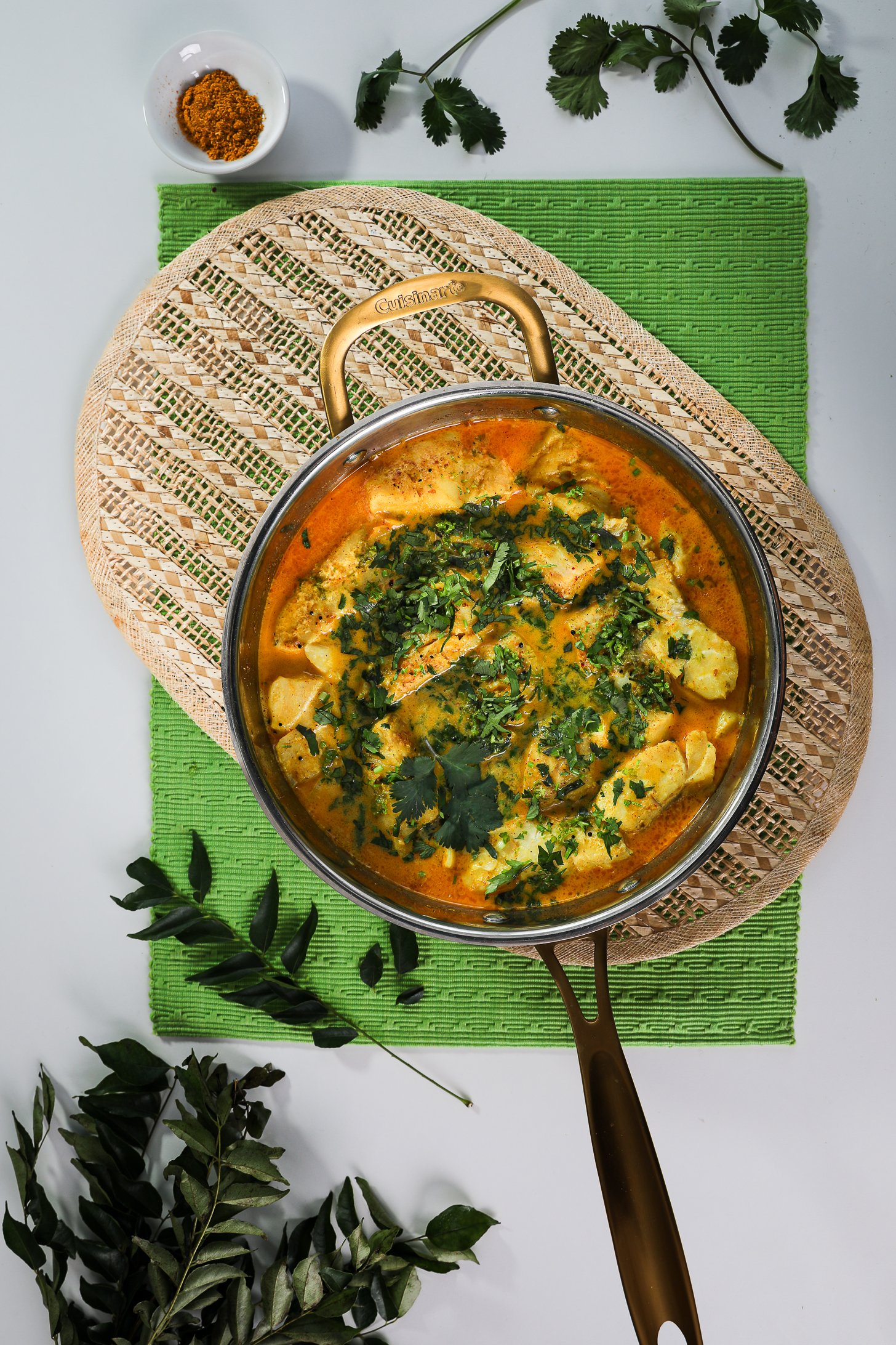Bird's-eye view: Pan of fish fillets immersed in vibrant curry sauce, topped with chopped cilantro, elegantly styled with mats and curry leaves.