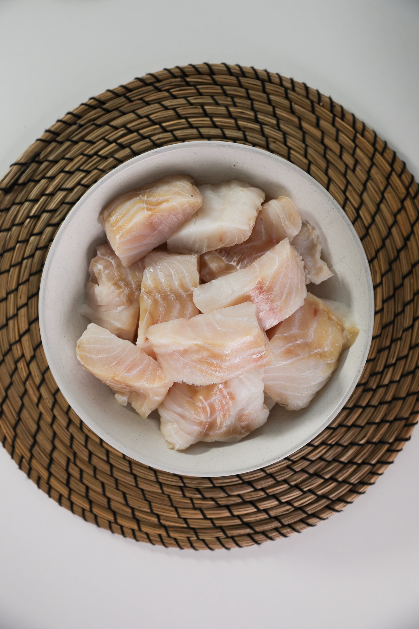 A bowl of medium-sized cod fish fillets placed on a round straw mat.