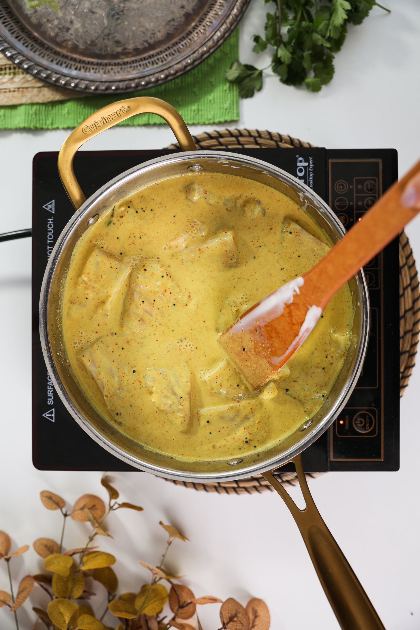 Mobile cooktop showcasing a pan of fish fillets gently simmering in curry sauce, accompanied by a wooden spatula.