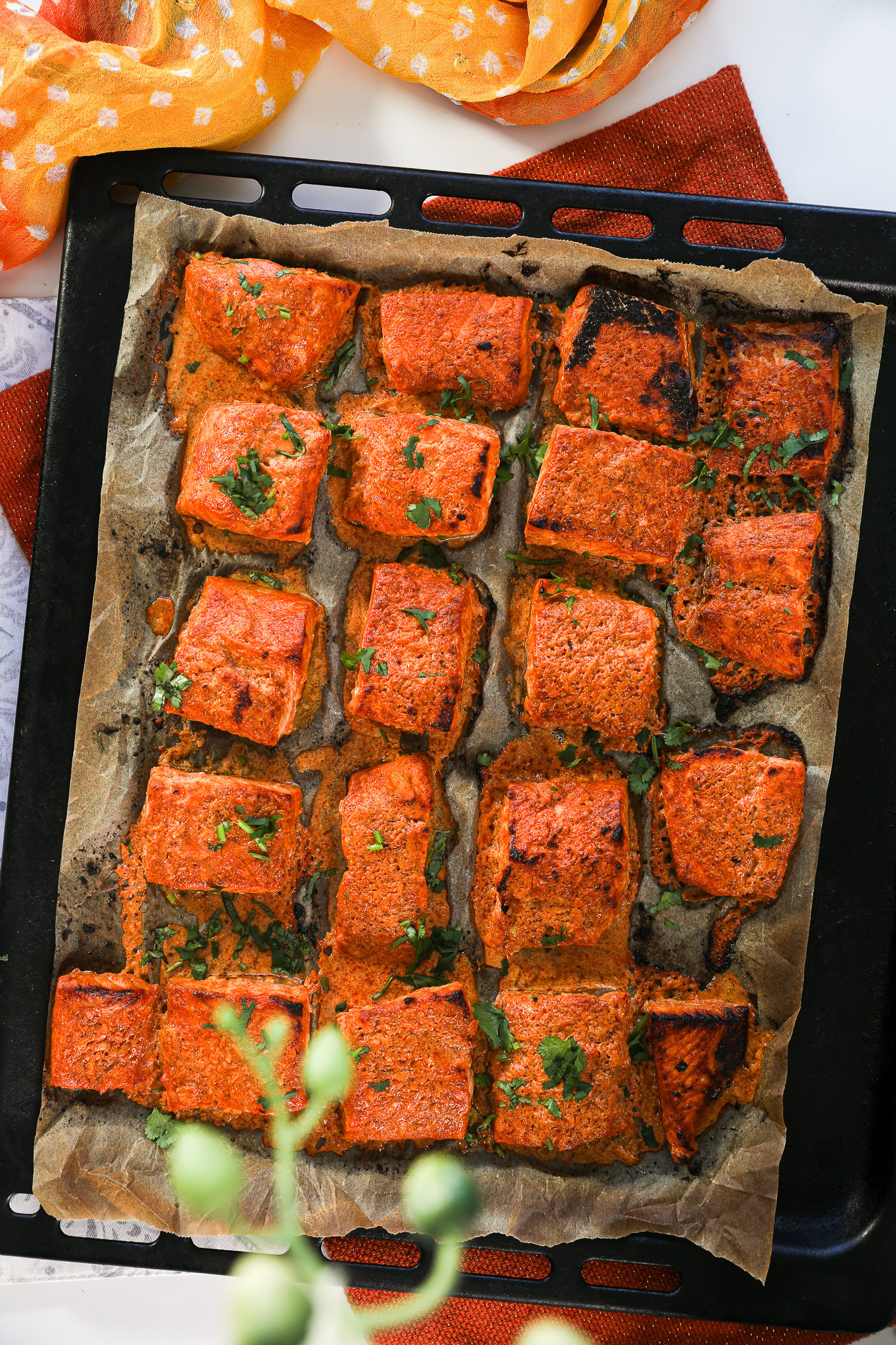 Overhead close up shot of a sheet pan featuring rows of salmon tandoori fish tikka, artfully styled on an orange mat with a traditional scarf.