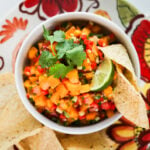 A close up top view image of a bowl of mango salsa topped with cilantro and lime alongside nachos.