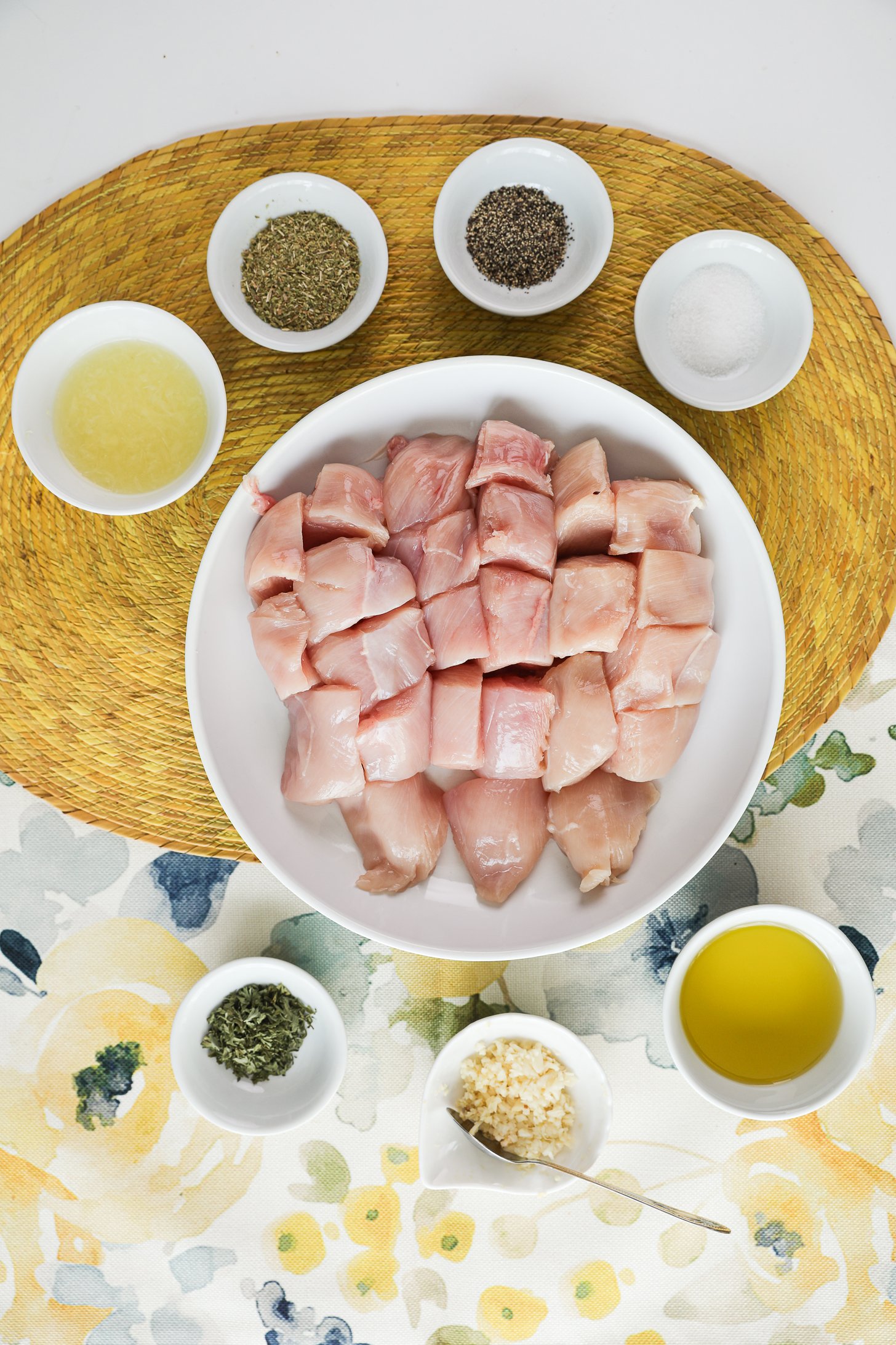 A selection of food ingredients including a bowl of raw chicken breast cubes, spices, oil, lemon and crushed garlic.