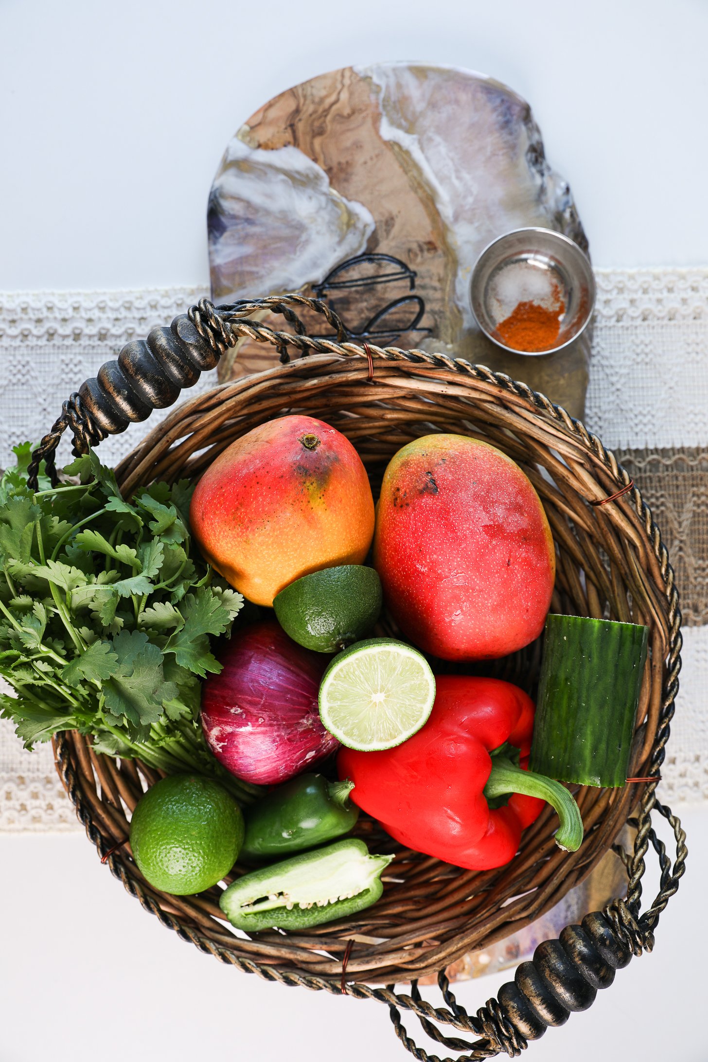 A basket of whole produce including mangoes, pepper, cucumber, jalapeno, onion, limes and cilantro.