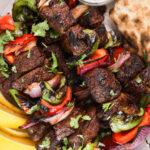 Zoomed-in image of a pile of grilled shish kabob skewers on a plate with a yogurt dip and lemon segments.