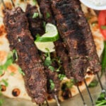 Close-up top-down view of beef kebabs on skewers atop a naan, decorated with a lime swirl and cilantro leaves and a ramekin of raita.