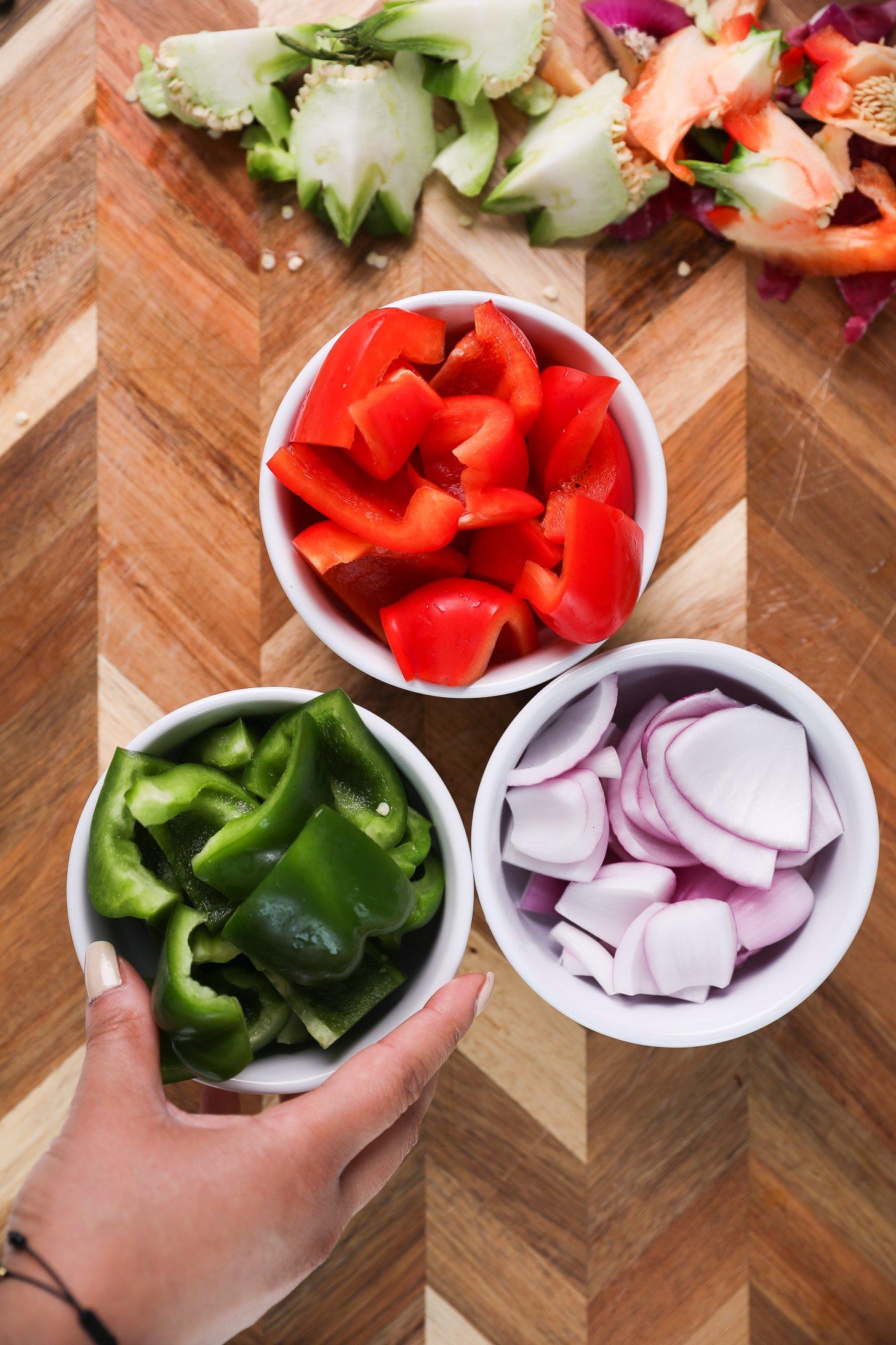 Three bowls of sliced vegetables: red and green bell pepper and red onion with a hand holding one of the bowls.
