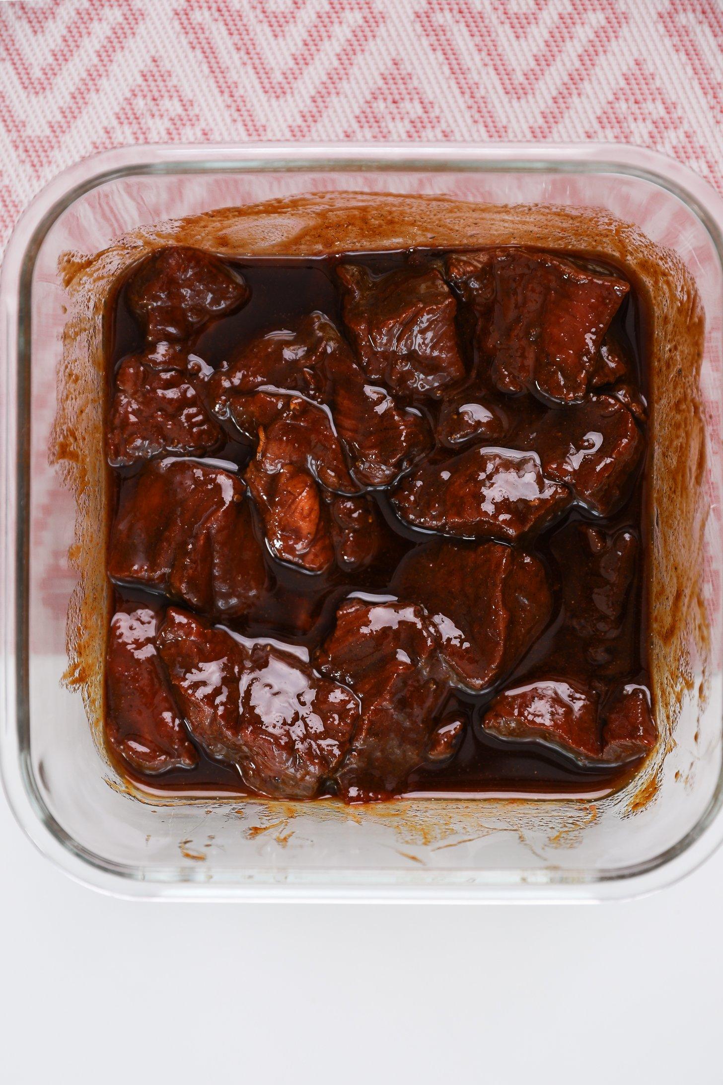 A glass container of beef cubes submerged in a dark brown marinade.