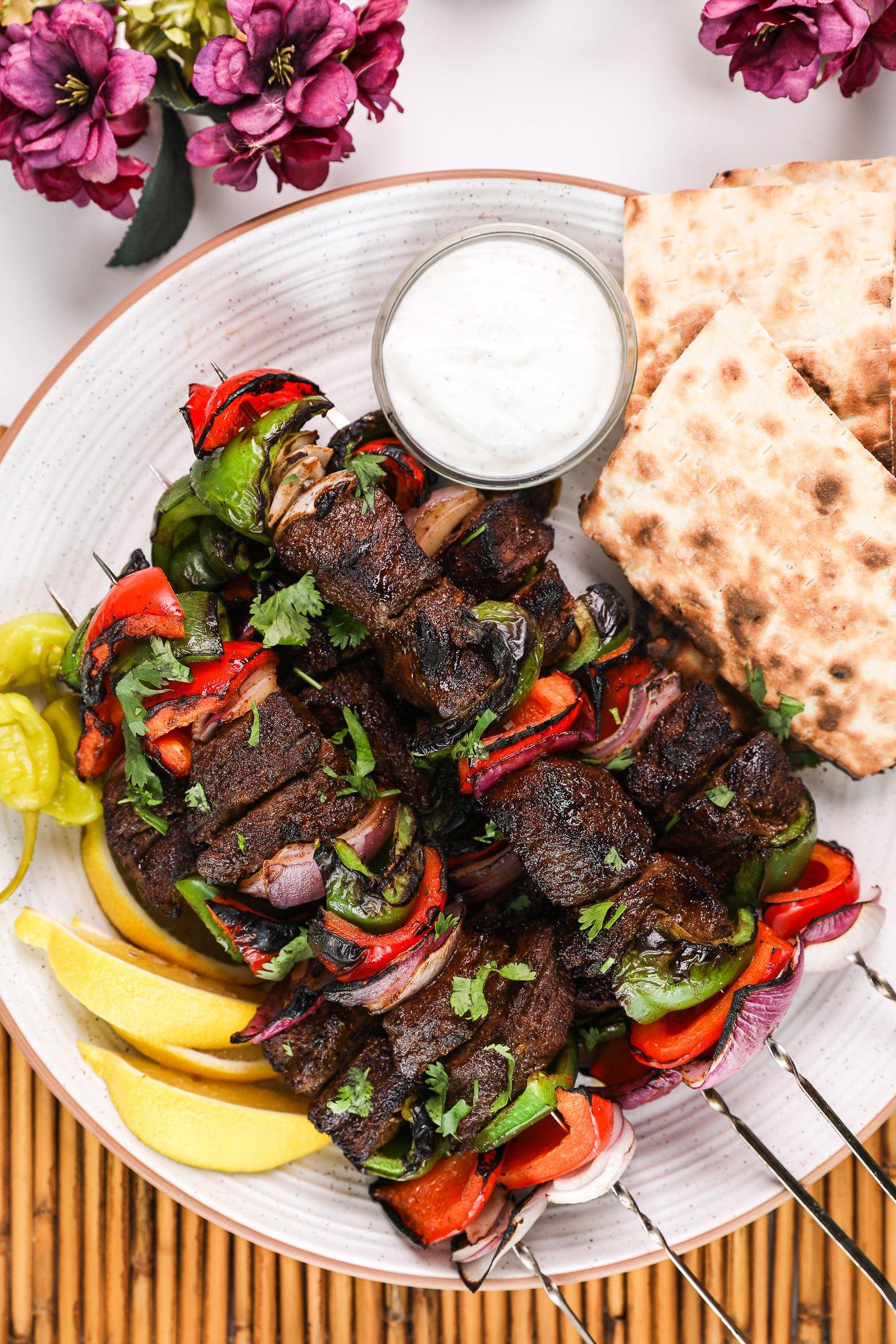 Overhead image of a plate of grilled shish kabob skewers and peppers alongside a yogurt dip and pieces of pita bread.