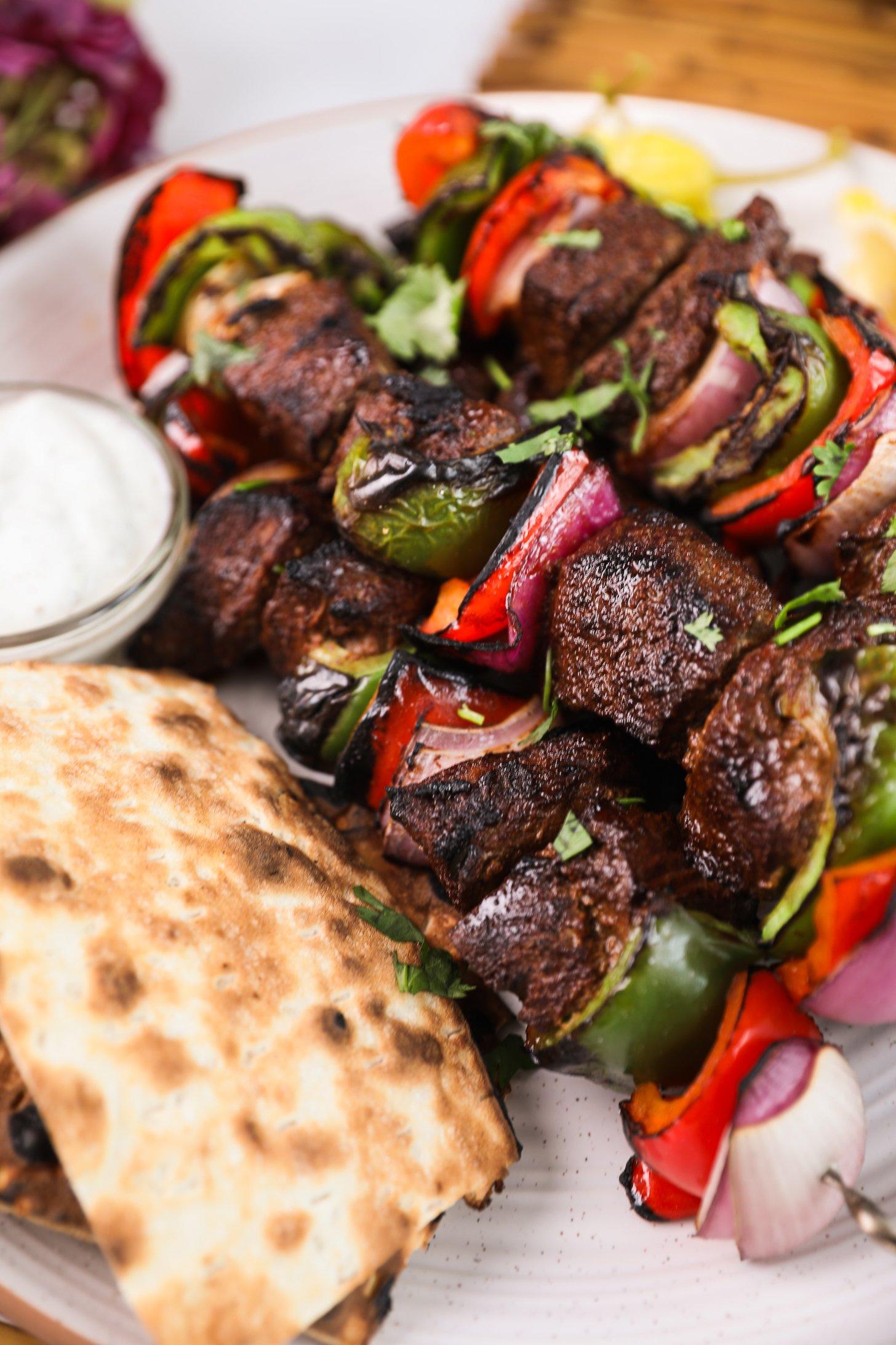 A perspective image of grilled shish kabob skewers with peppers alongside pita bread and a yogurt dip.