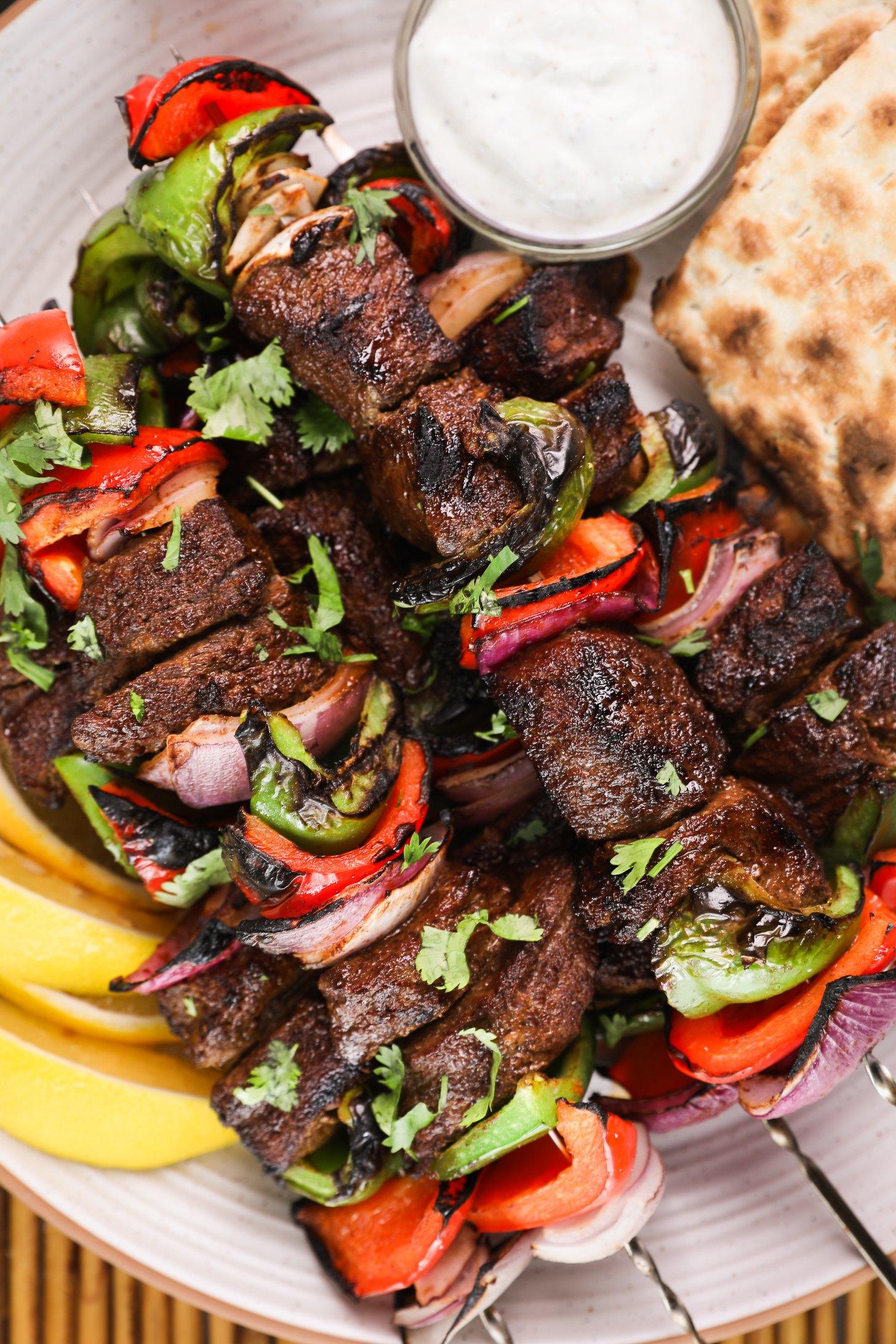 Zoomed-in image of a pile of grilled shish kabob skewers on a plate with a yogurt dip and lemon segments.