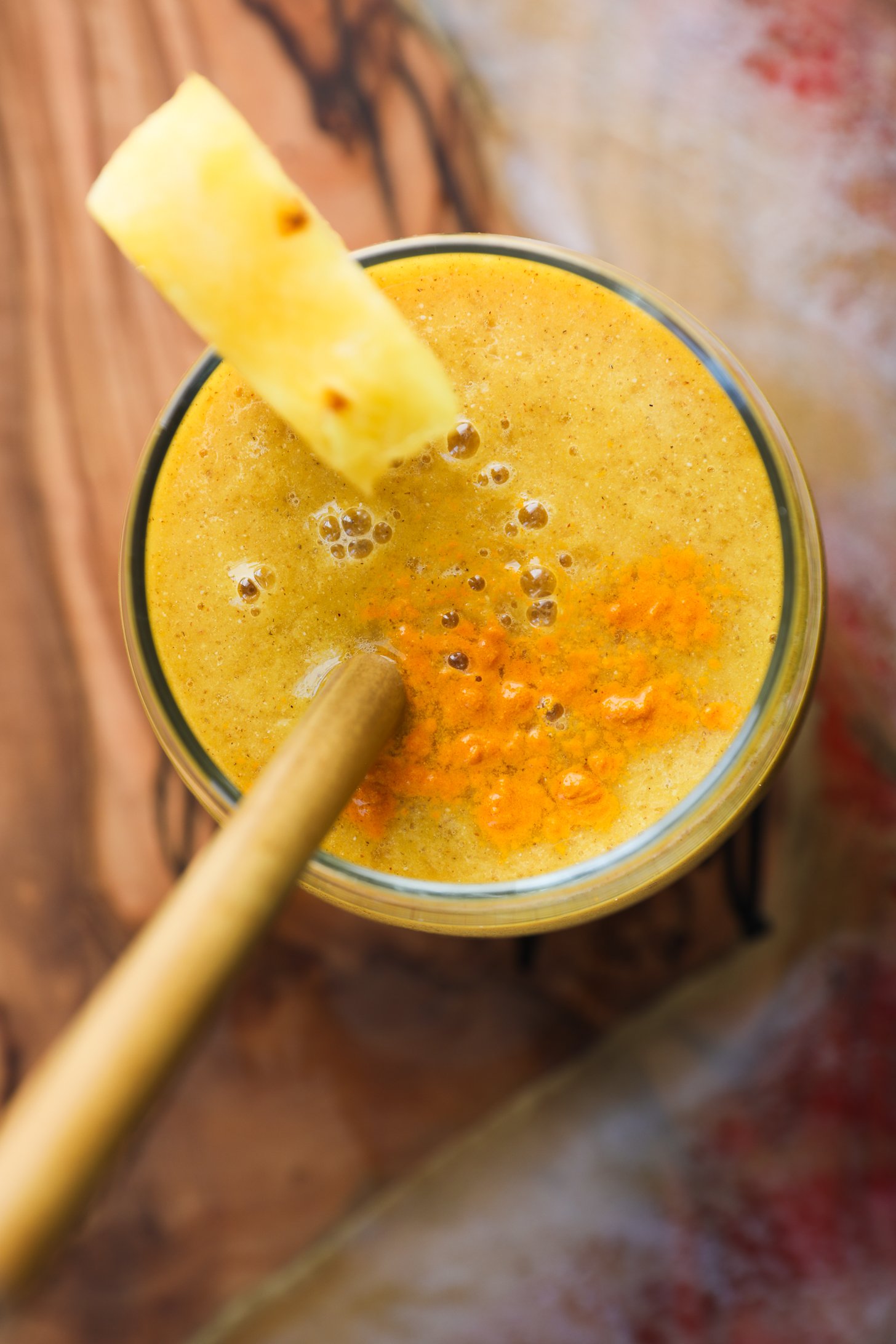 Top view of a yellow smoothie sprinkled with turmeric powder, garnished with a pineapple wedge on the rim, and a wooden straw nestled inside.