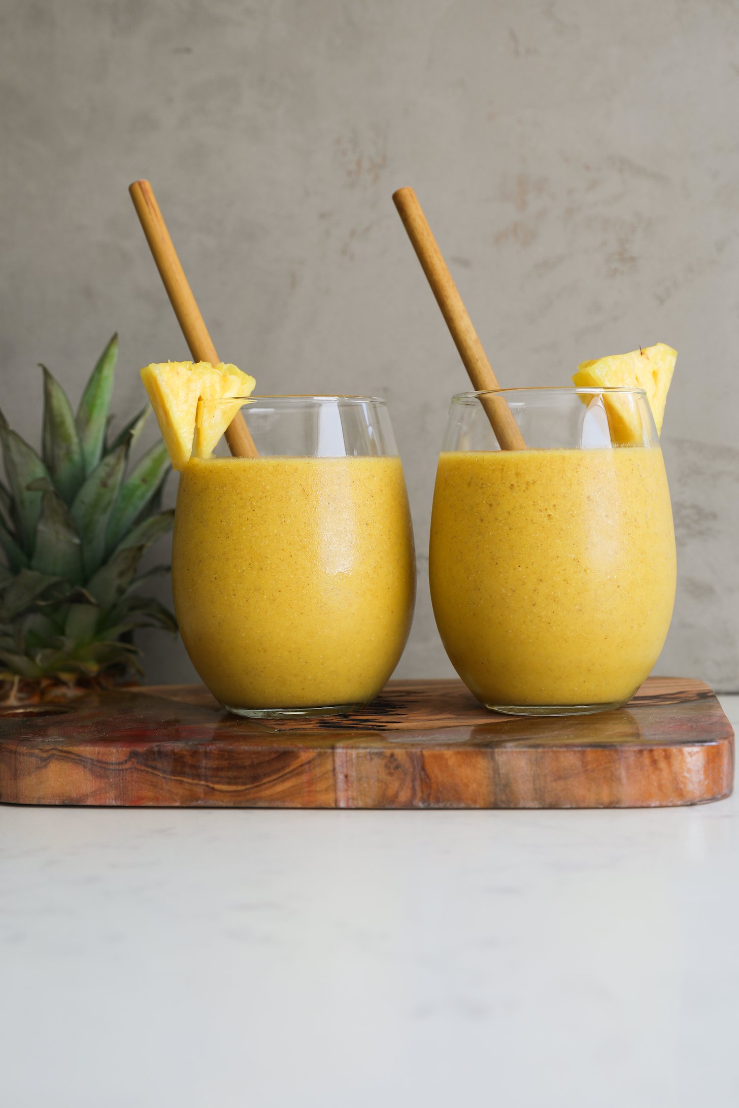 Perspective image of two glasses of yellow smoothie with bamboo straws in them.